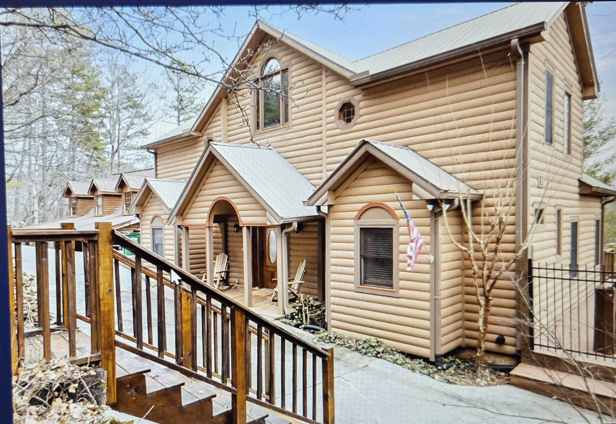 A wooden two-story house with a porch, stairs, and trees surrounding it in a winter setting.