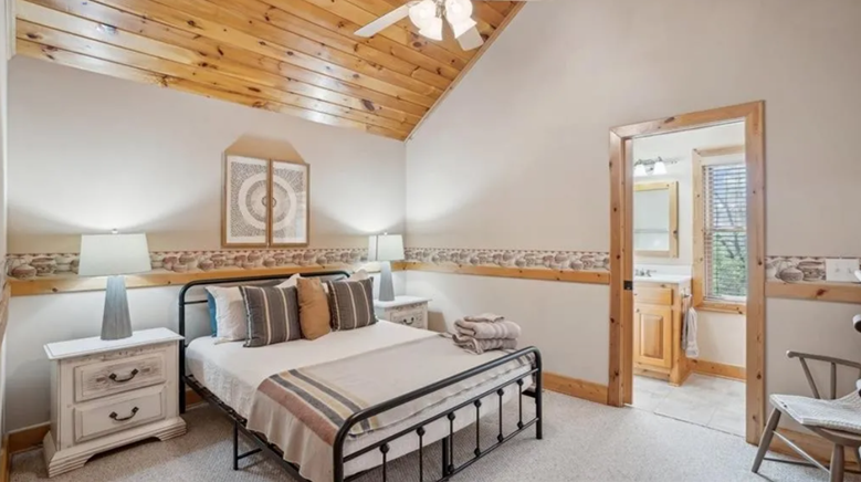 A cozy bedroom with a wooden ceiling, a metal bed frame, and two white nightstands with lamps. The bed has multiple pillows and a folded blanket at the foot. There's a decorative piece of art above the headboard, and a doorway leading to a small bath