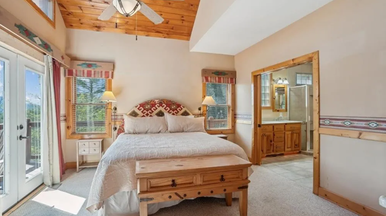 Cozy bedroom with a white bed, wooden furniture, windows, and a view of greenery outside.
