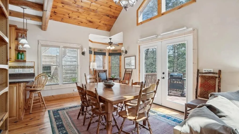 Cozy dining area in a cabin-style living room with wooden ceiling, large windows, and glass door leading to outdoor deck.