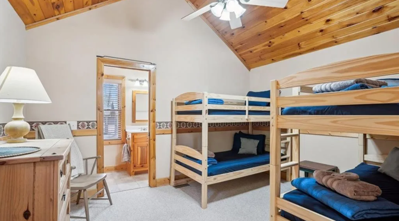 A bedroom with two wooden bunk beds, a wooden dresser with a lamp, and a small chair. The room has a wooden ceiling, white walls with decorative border, and an open door leading to a bathroom.