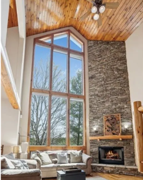 Interior of a living room with a tall, large window, a stone fireplace, a sectional sofa, and a wooden ceiling.