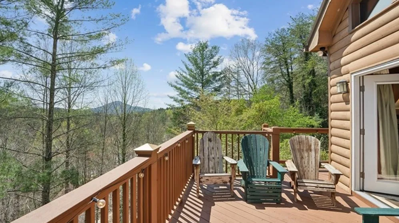 A wooden deck with three Adirondack chairs overlooking a forested landscape with trees and distant mountains under a partly cloudy sky.
