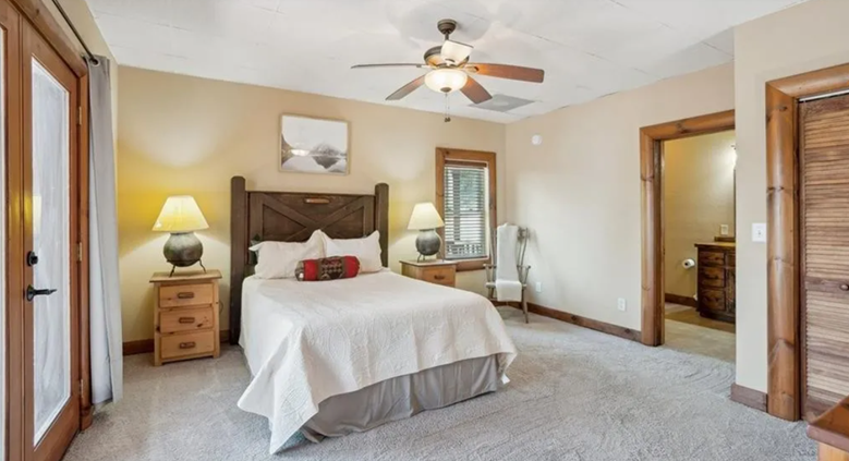 Bedroom with a large bed, wooden headboard, white bedding, two nightstands with lamps, a chair, a ceiling fan, and a window with blinds.