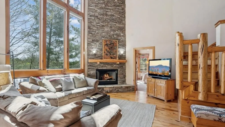 Living room with large windows, stone fireplace, beige sofa, wooden staircase, TV, and wood flooring.