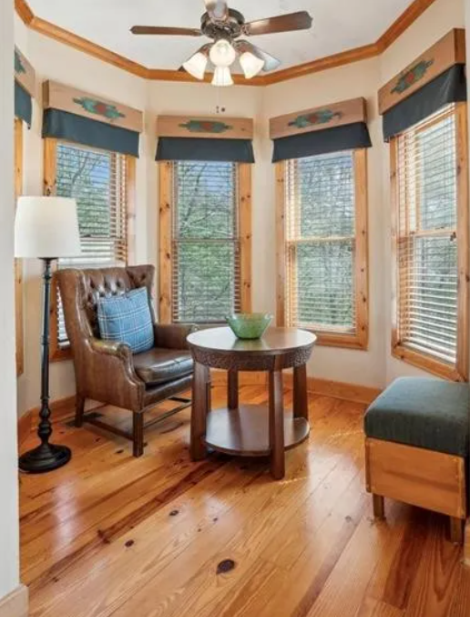 Sunlit corner with wooden flooring, a leather armchair with a plaid pillow, a round wooden table with a green bowl, a floor lamp, and multiple windows with blue valances and blinds.