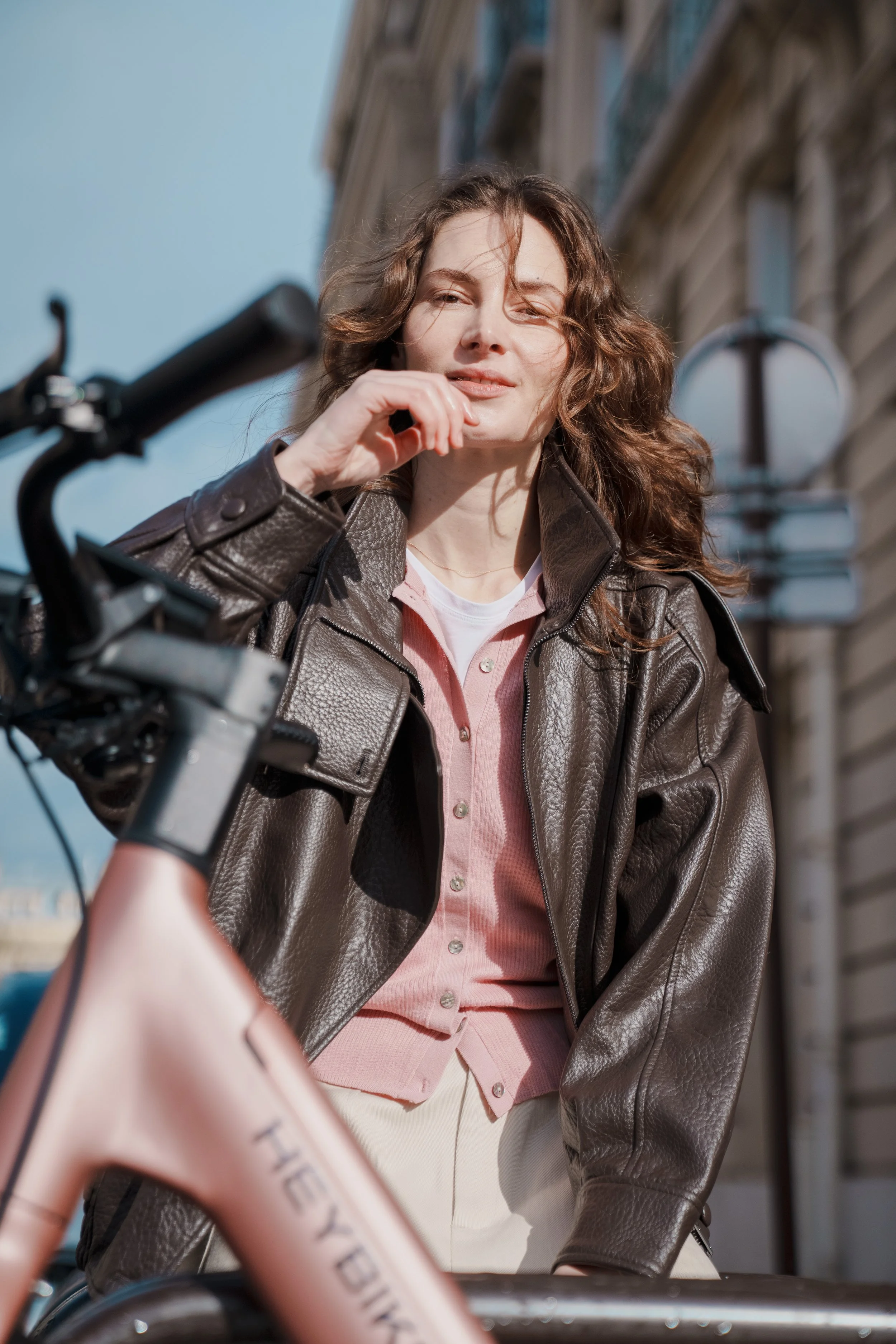 A woman with curly brown hair wearing a brown leather jacket and pink shirt, standing outdoors with a bicycle and architectural buildings in the background.