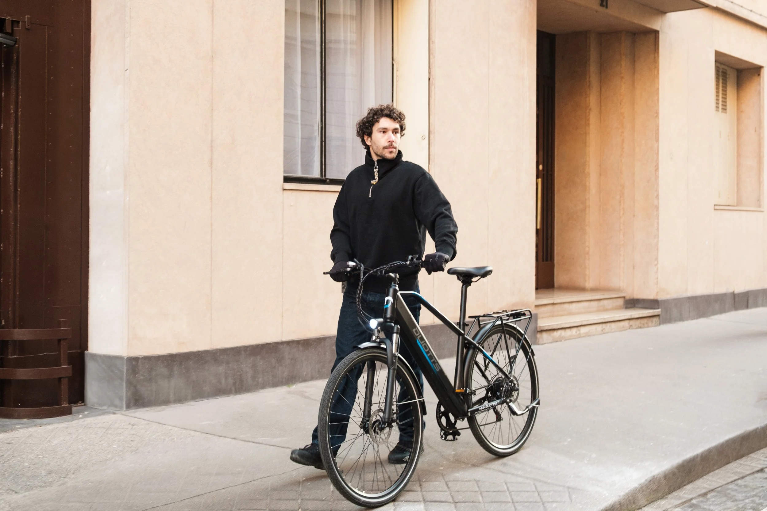 A man standing with a black bicycle on a city sidewalk in front of a beige building with large windows and a doorway.