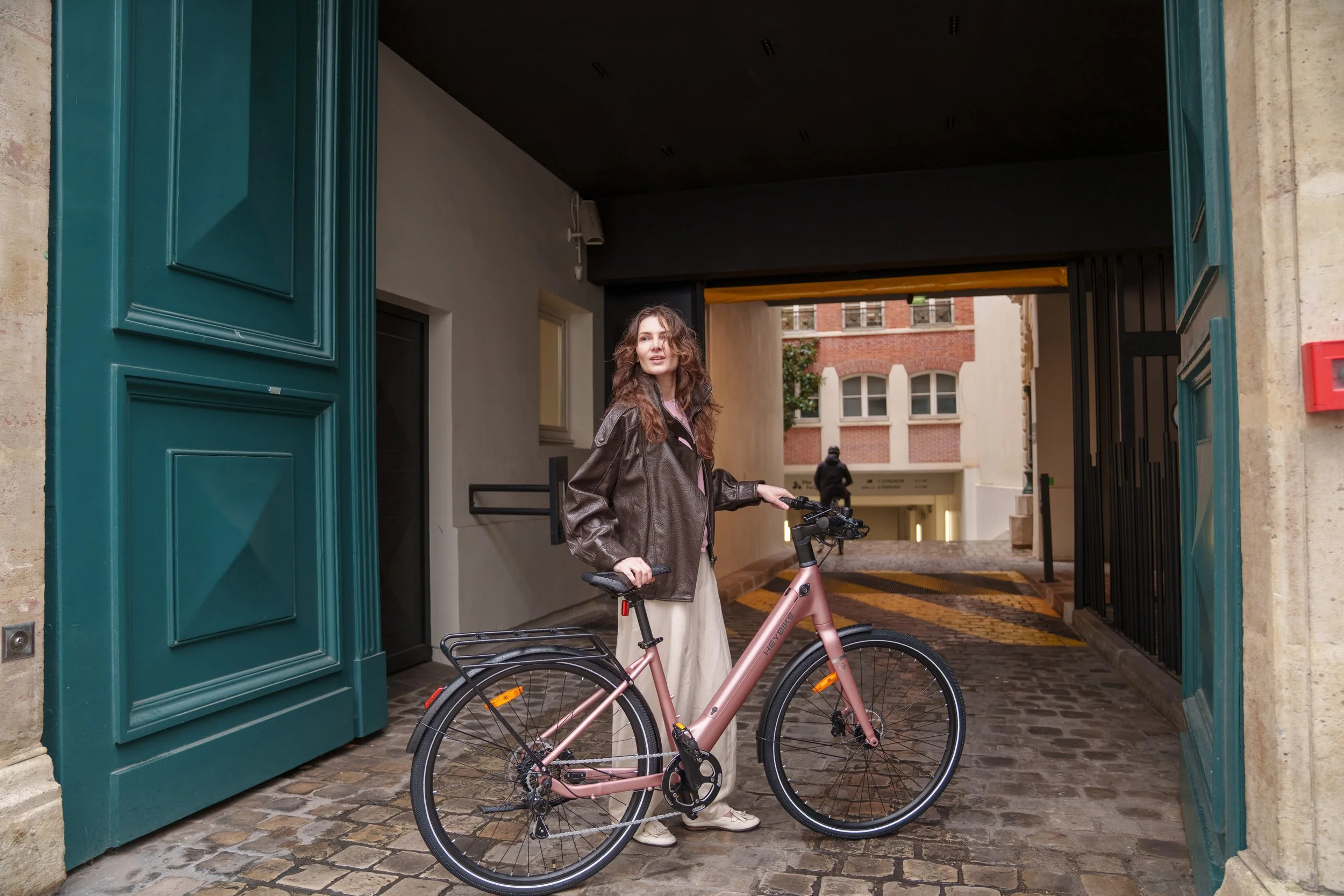 A woman in a brown leather jacket and beige pants standing with a pink bicycle in a cobblestone alleyway near a green door.