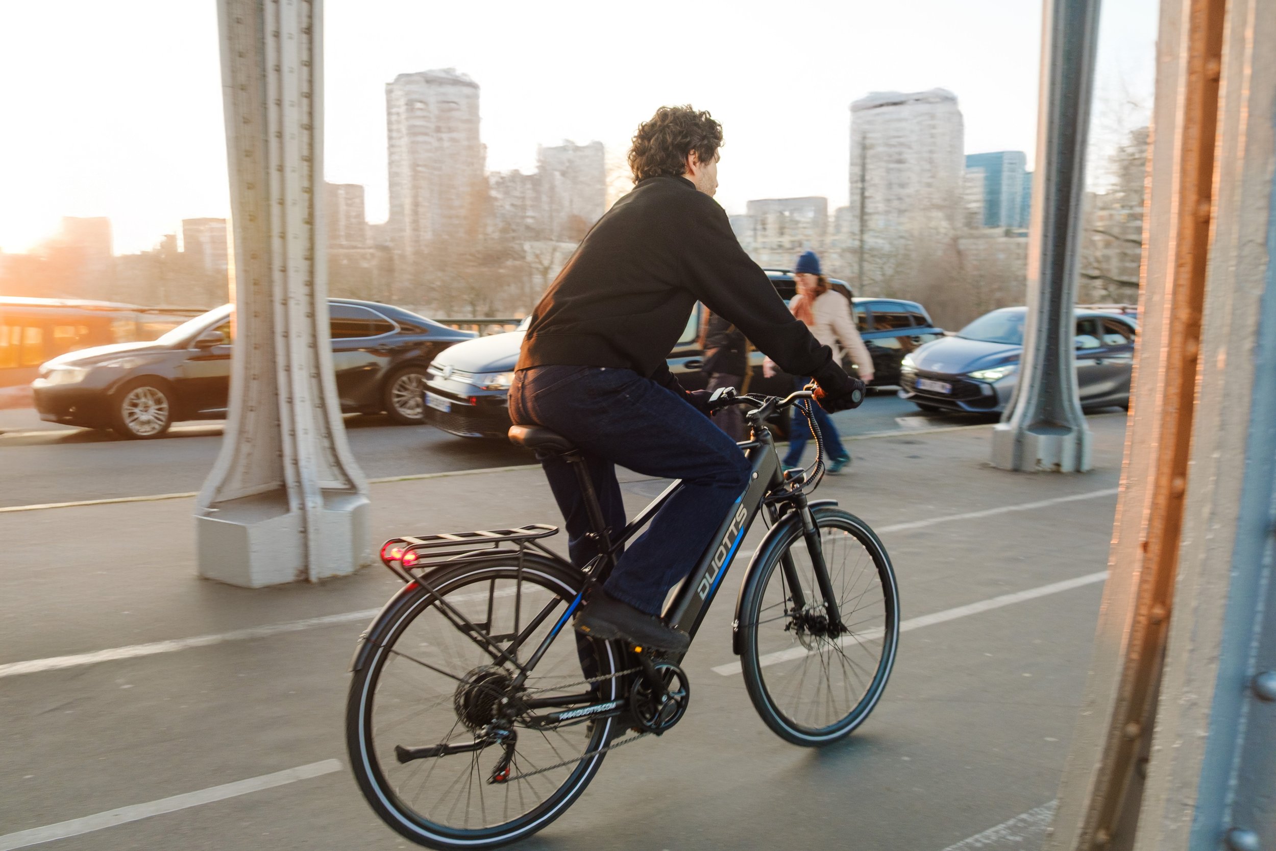 A person riding a black electric bicycle on a city street during sunset, with cars and pedestrians in the background.
