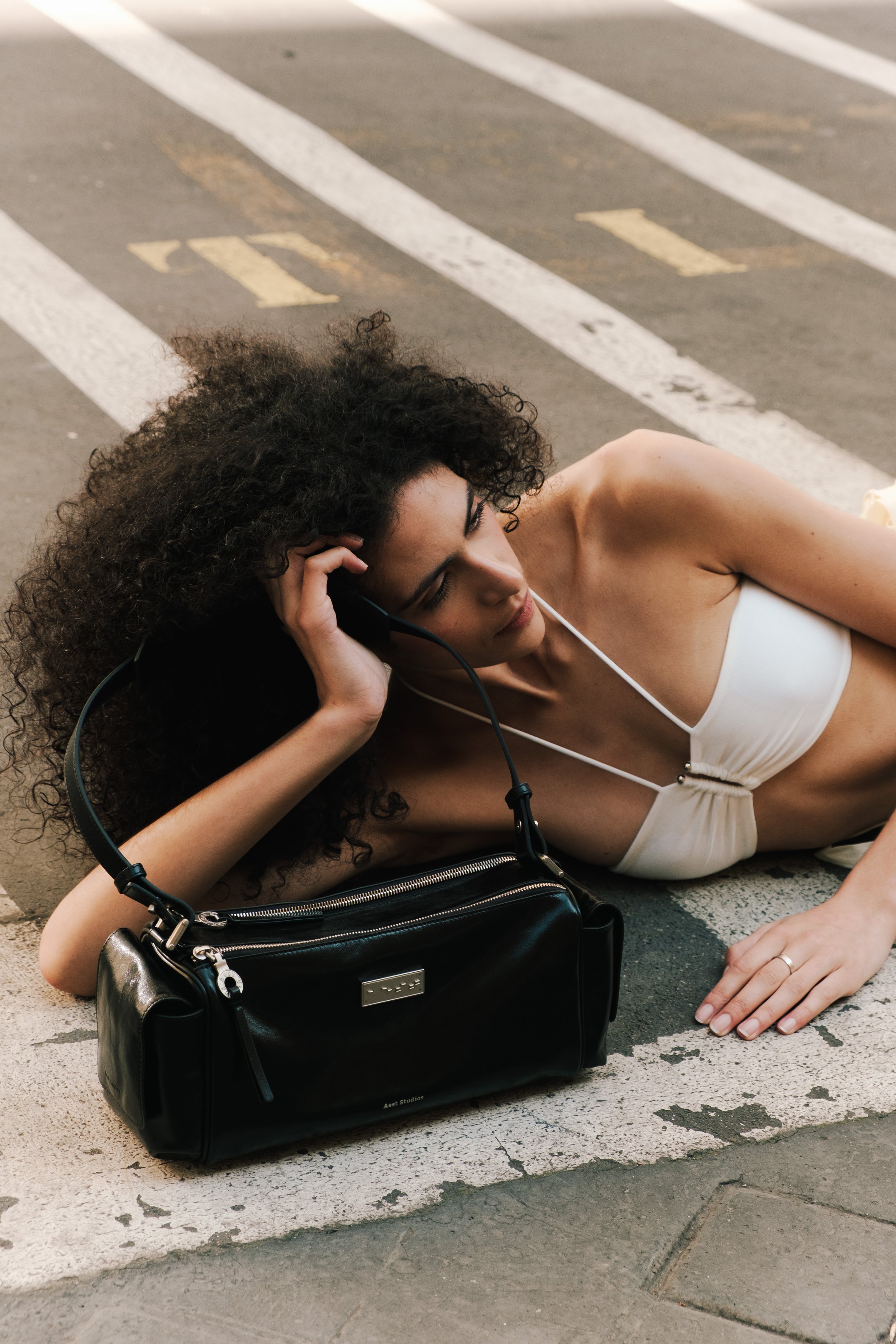A woman lying on the ground in a parking lot, wearing a white bikini top, with headphones on, and an expression of contemplation.