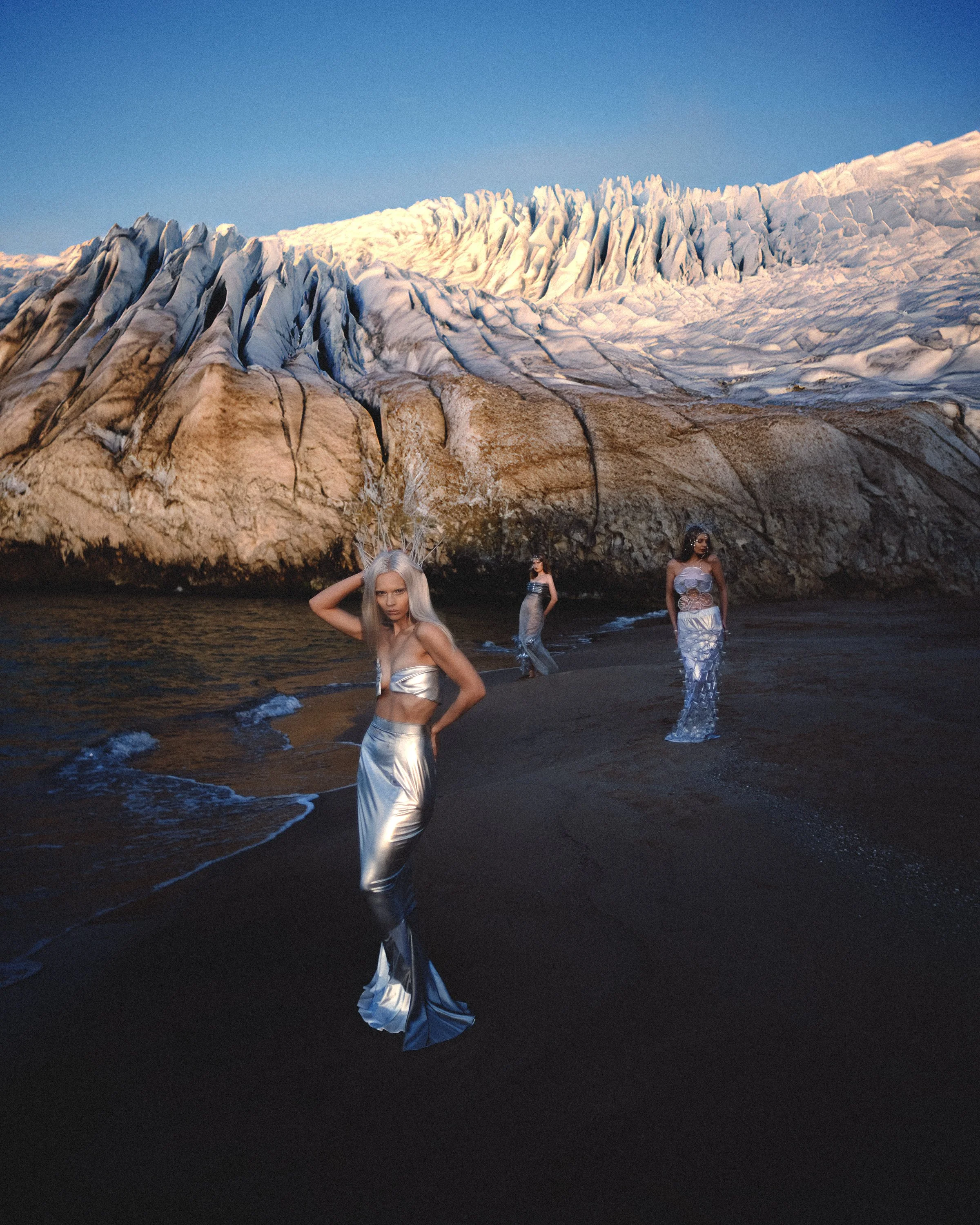 Three women in metallic clothing standing on a black sand beach near large ice formations and a glacier.
