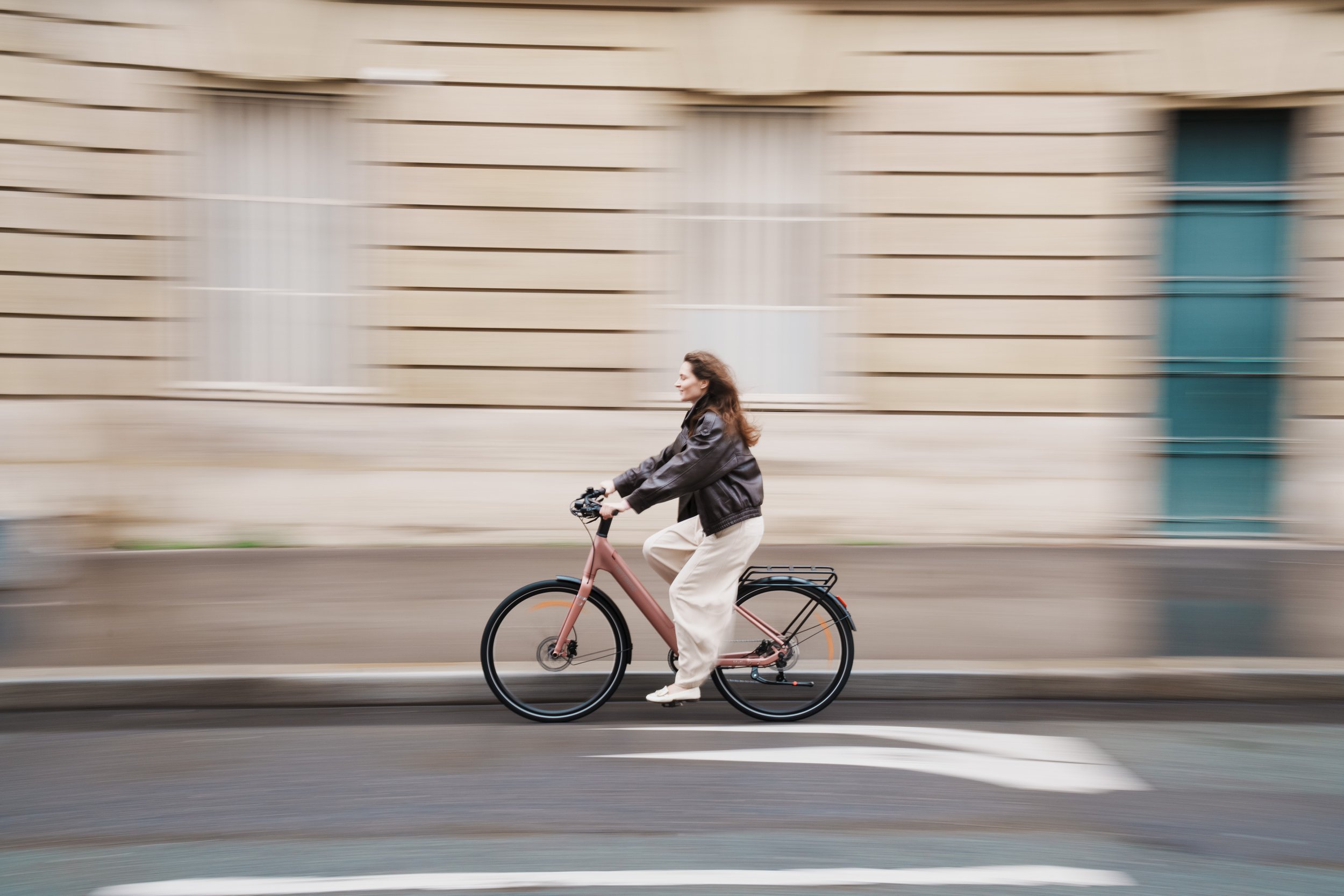 Woman riding a pink bicycle on a city street with a blurred beige building in the background.