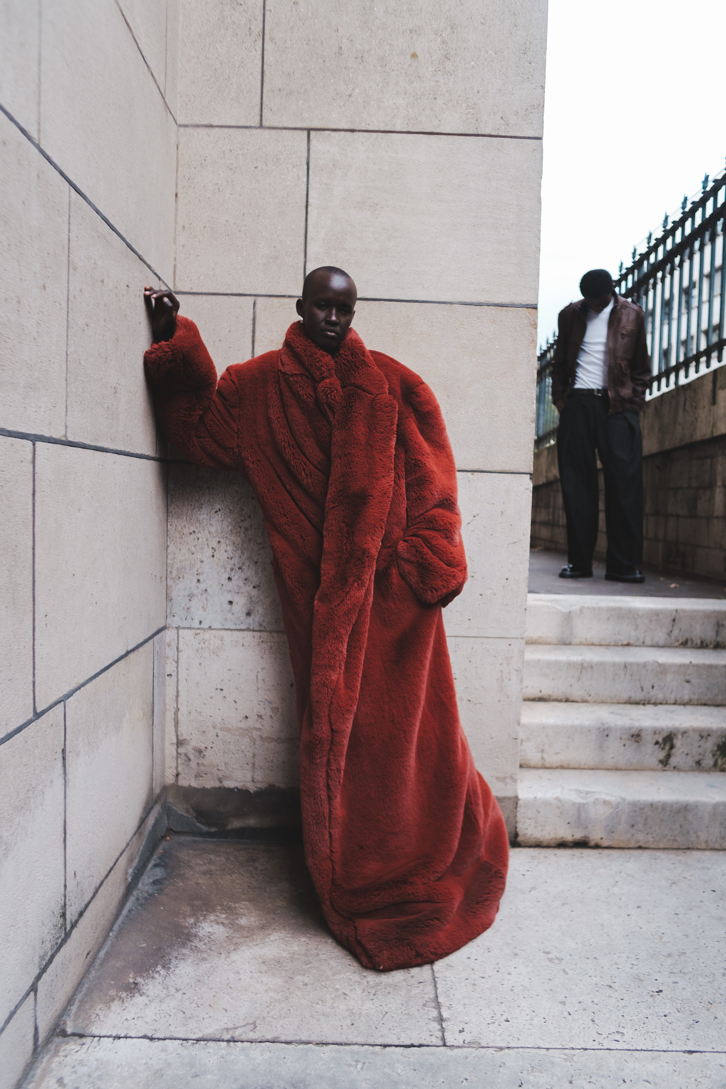 Person in a long red fur coat leaning against a stone wall, with another person standing on stairs in the background.
