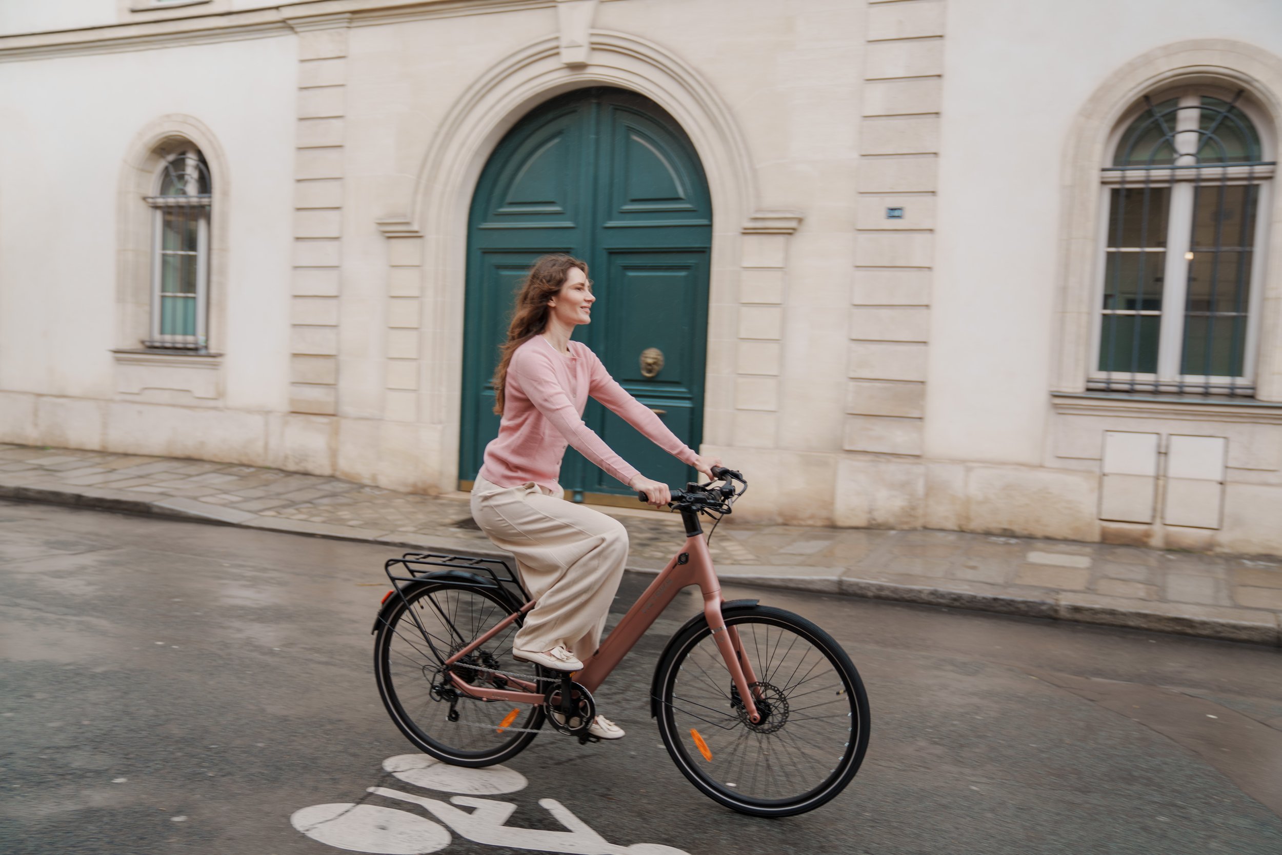 A woman riding a pink electric bike on a city street with cobblestone and pavement, in front of a white building with arched green door and large windows.