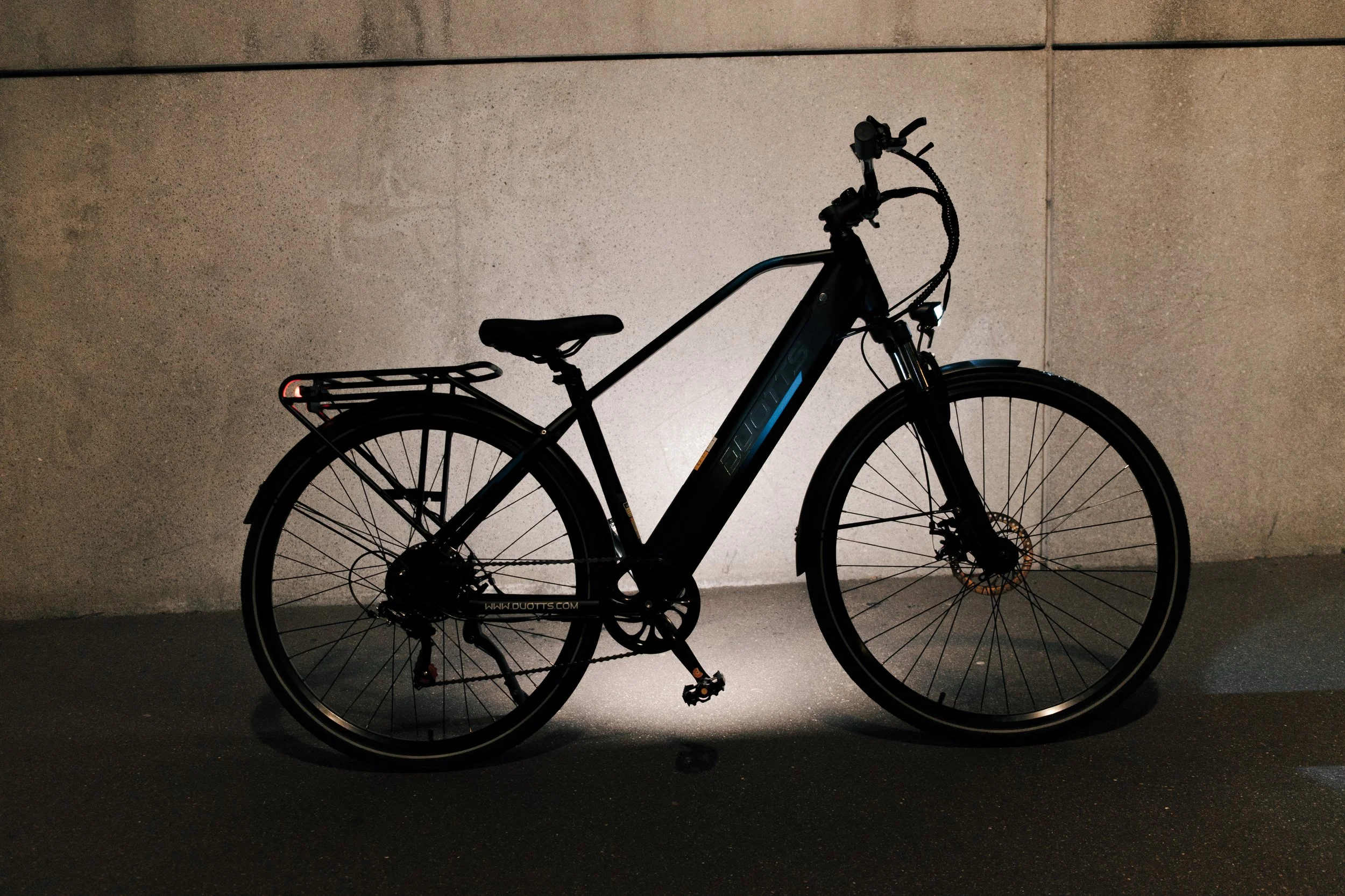 A black electric bike with a rear cargo rack, parked against a beige wall in a dimly lit setting.