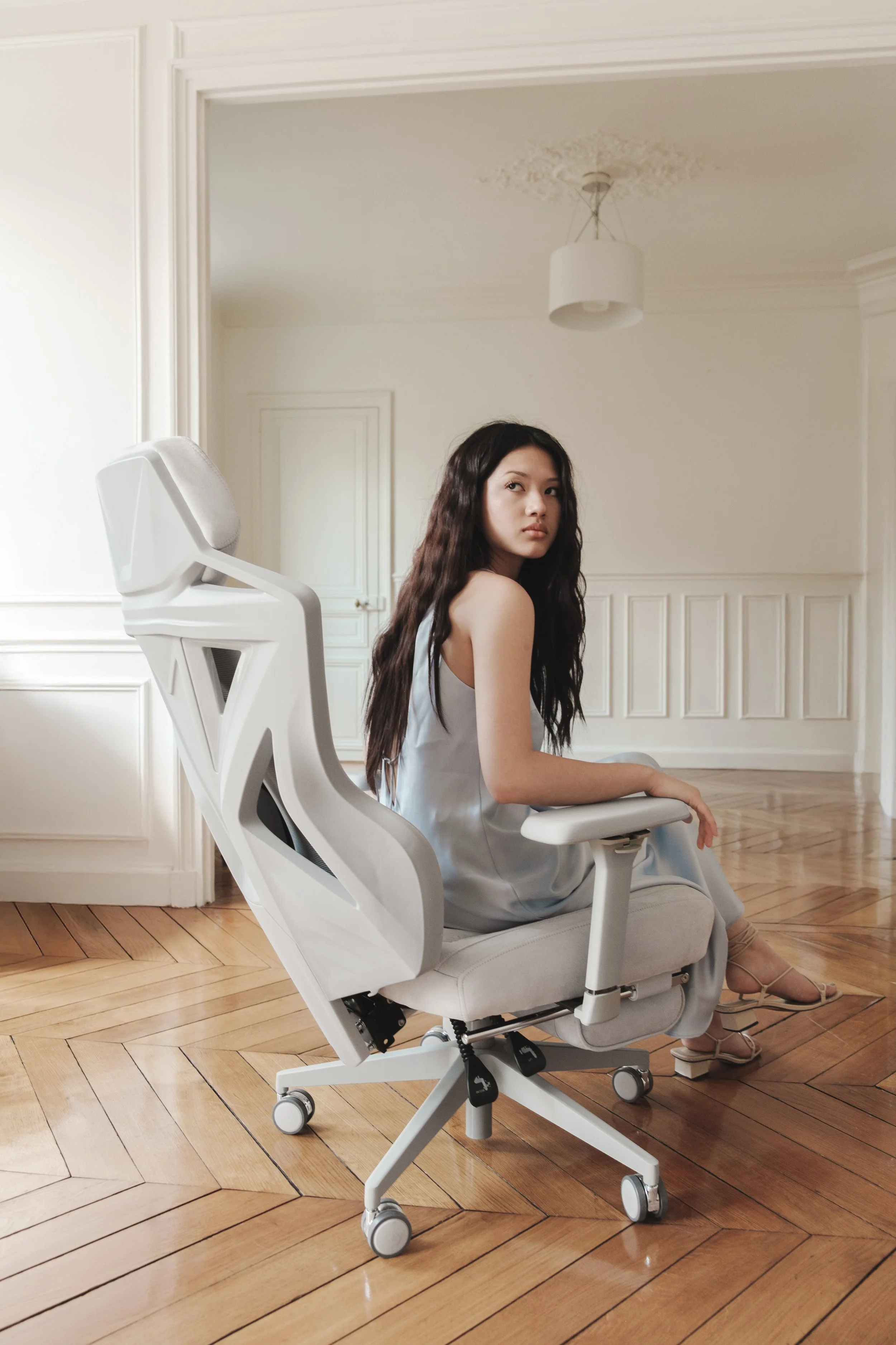 A woman sitting on a white ergonomic office chair in a room with wooden floors and white walls.