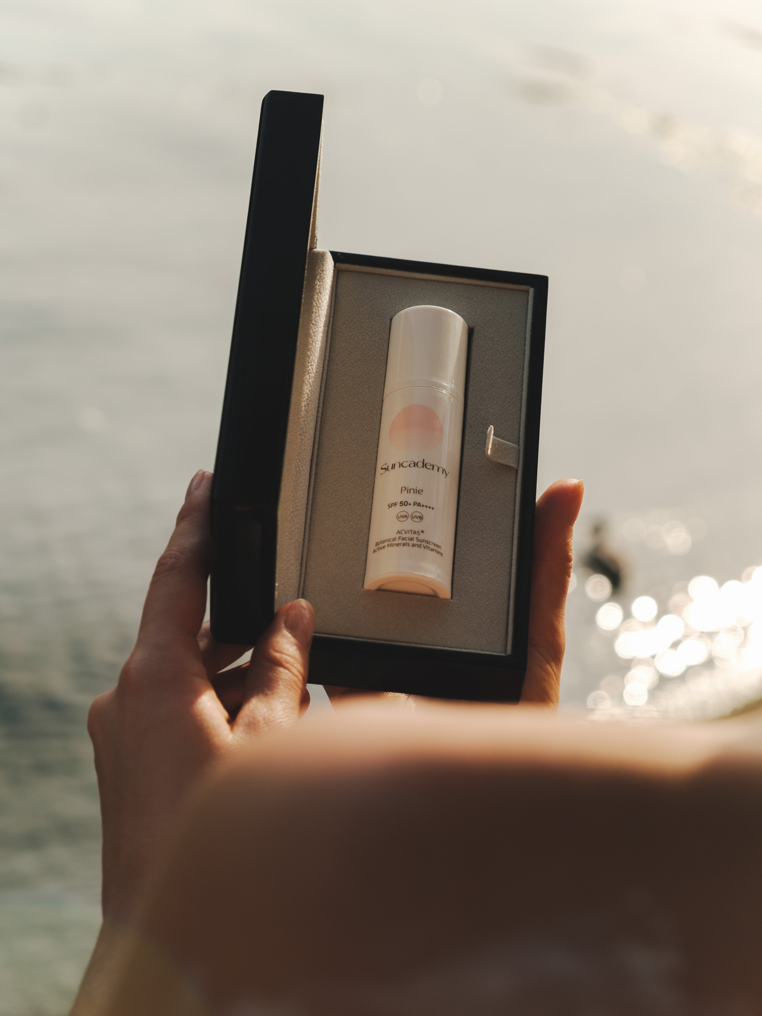 A hand holding a black box with a white sunscreen bottle inside, labeled 'Suncadamy' and 'Pinie', with a sunset or sunrise over the ocean in the background.