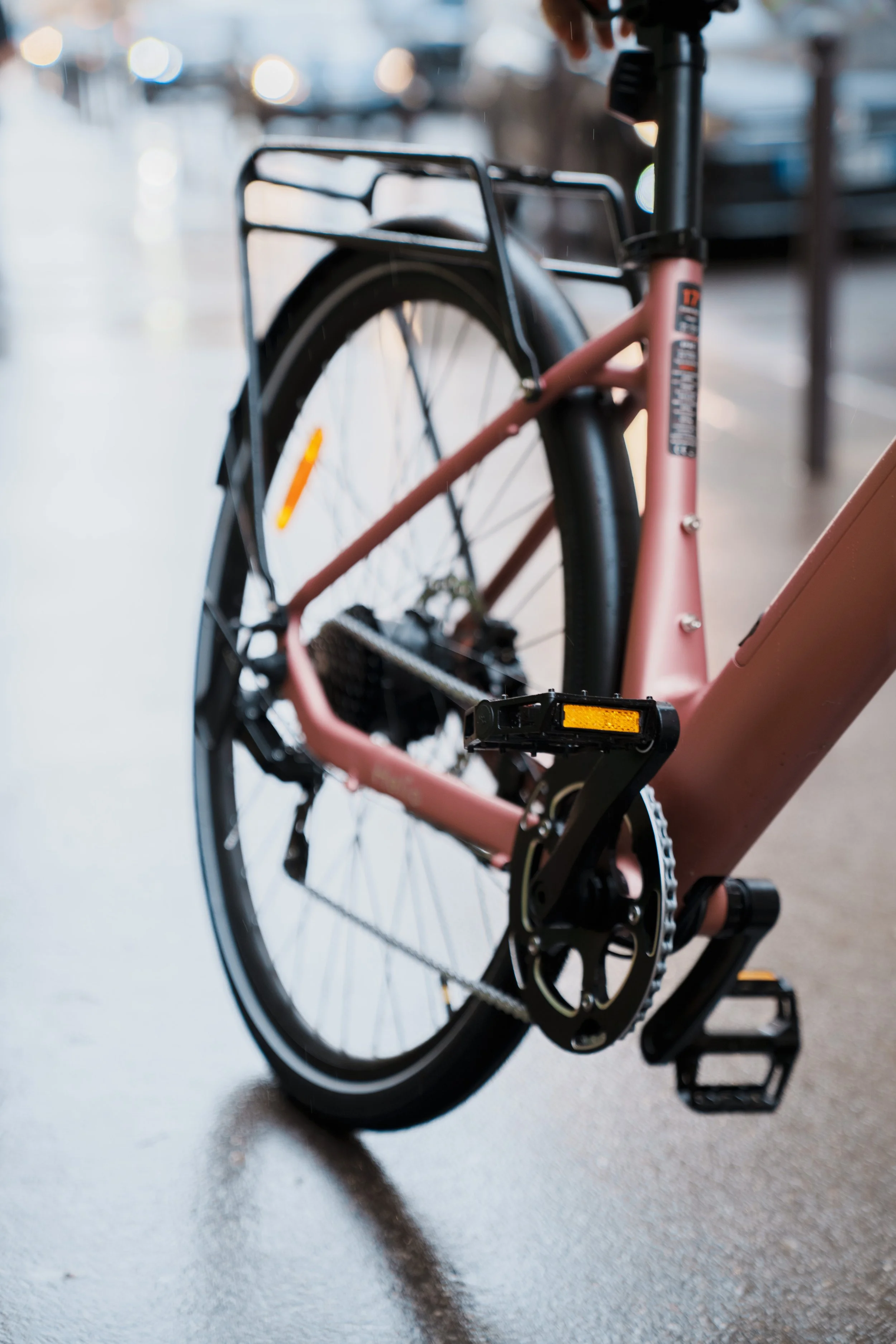 Close-up of a pink electric bicycle with black accents, showing the rear wheel, pedals, and frame, parked on a concrete surface with blurry background.