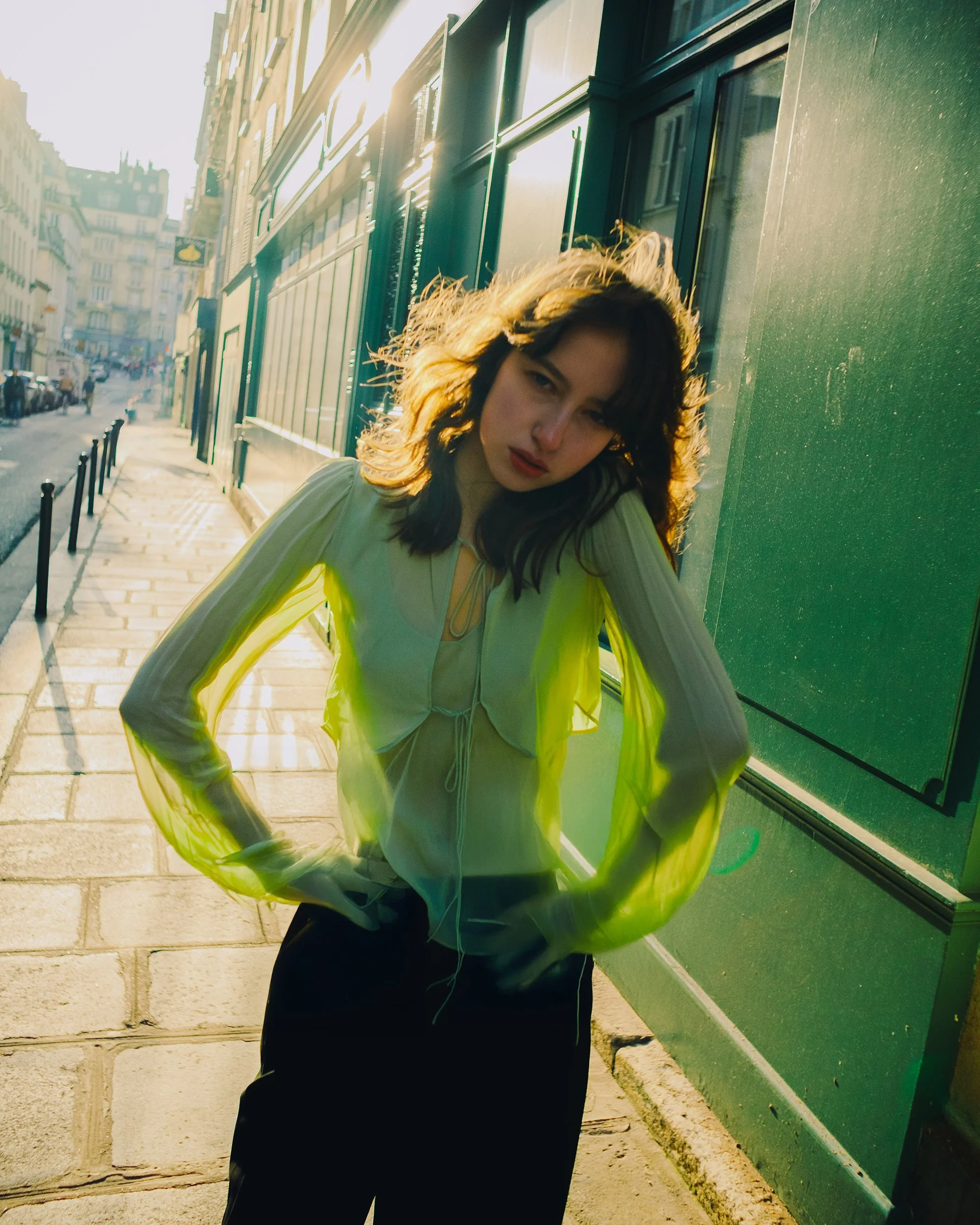 A young woman with wavy brown hair standing on a city sidewalk during sunset, wearing a white blouse with sheer sleeves and black pants, leaning against a green storefront window.