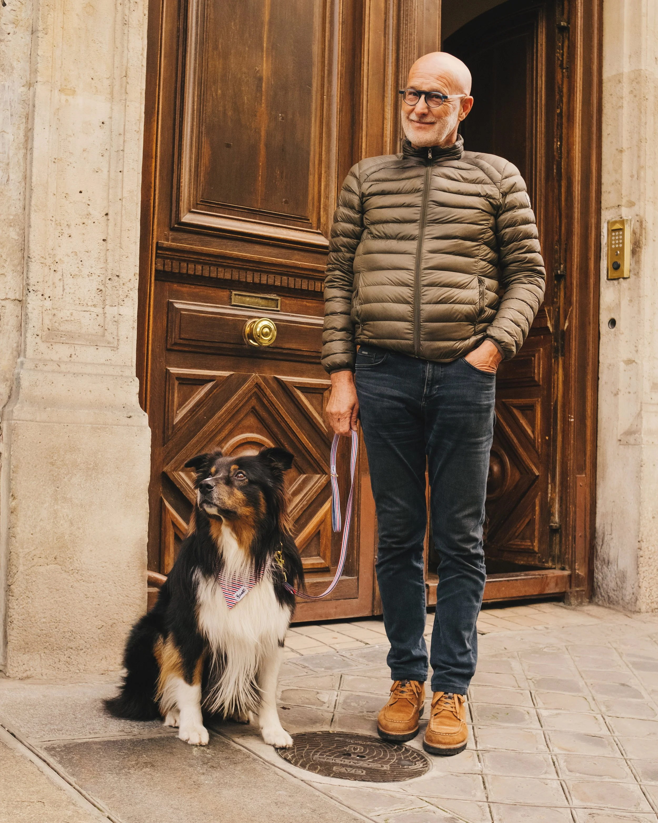 A man in a brown puffer jacket and jeans holding a leash with a black, white, and tan dog sitting next to him in front of a wooden door.
