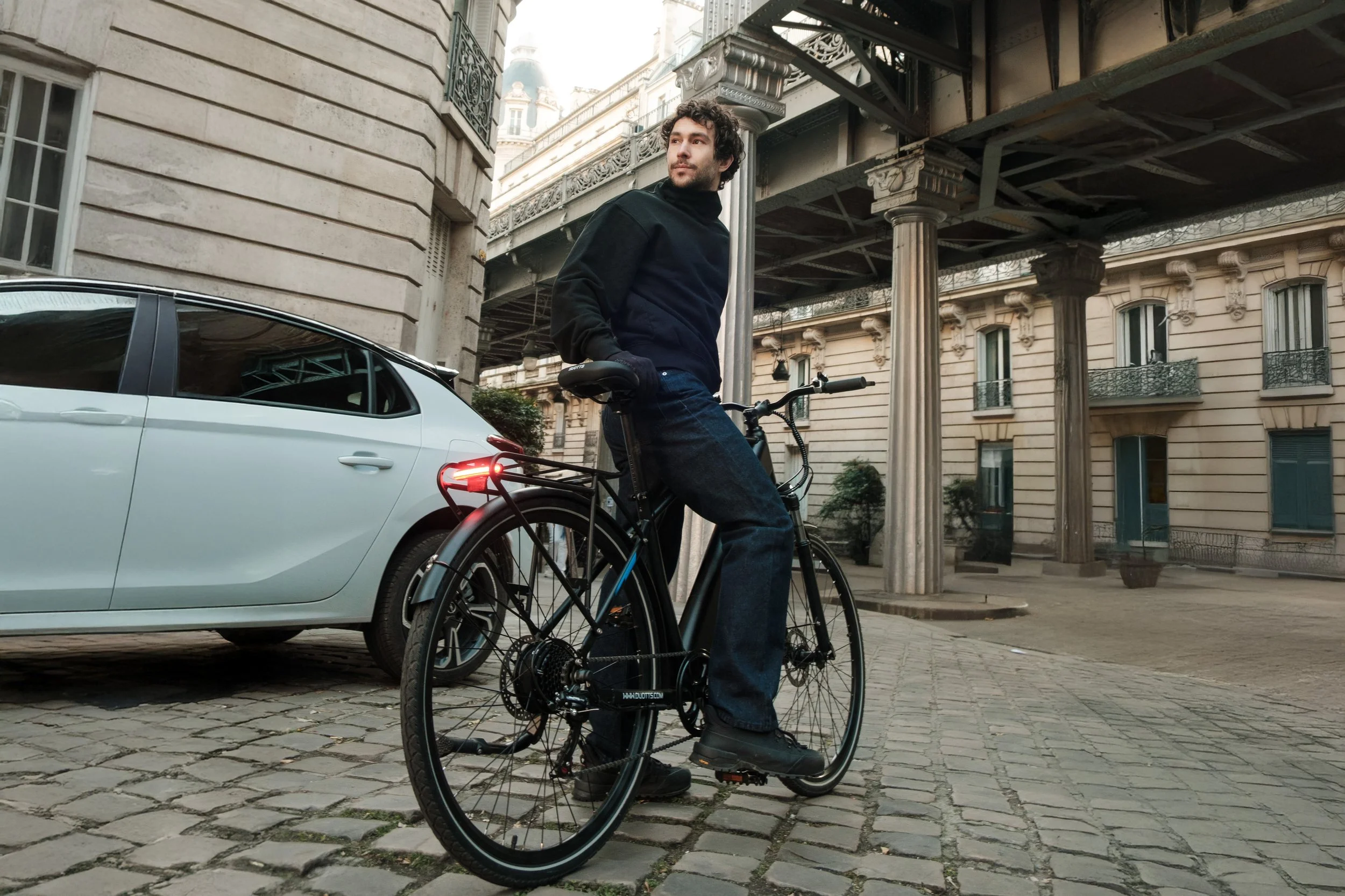 A man standing beside his black bicycle on a cobblestone street in front of a white car, with historic buildings and columns in the background.