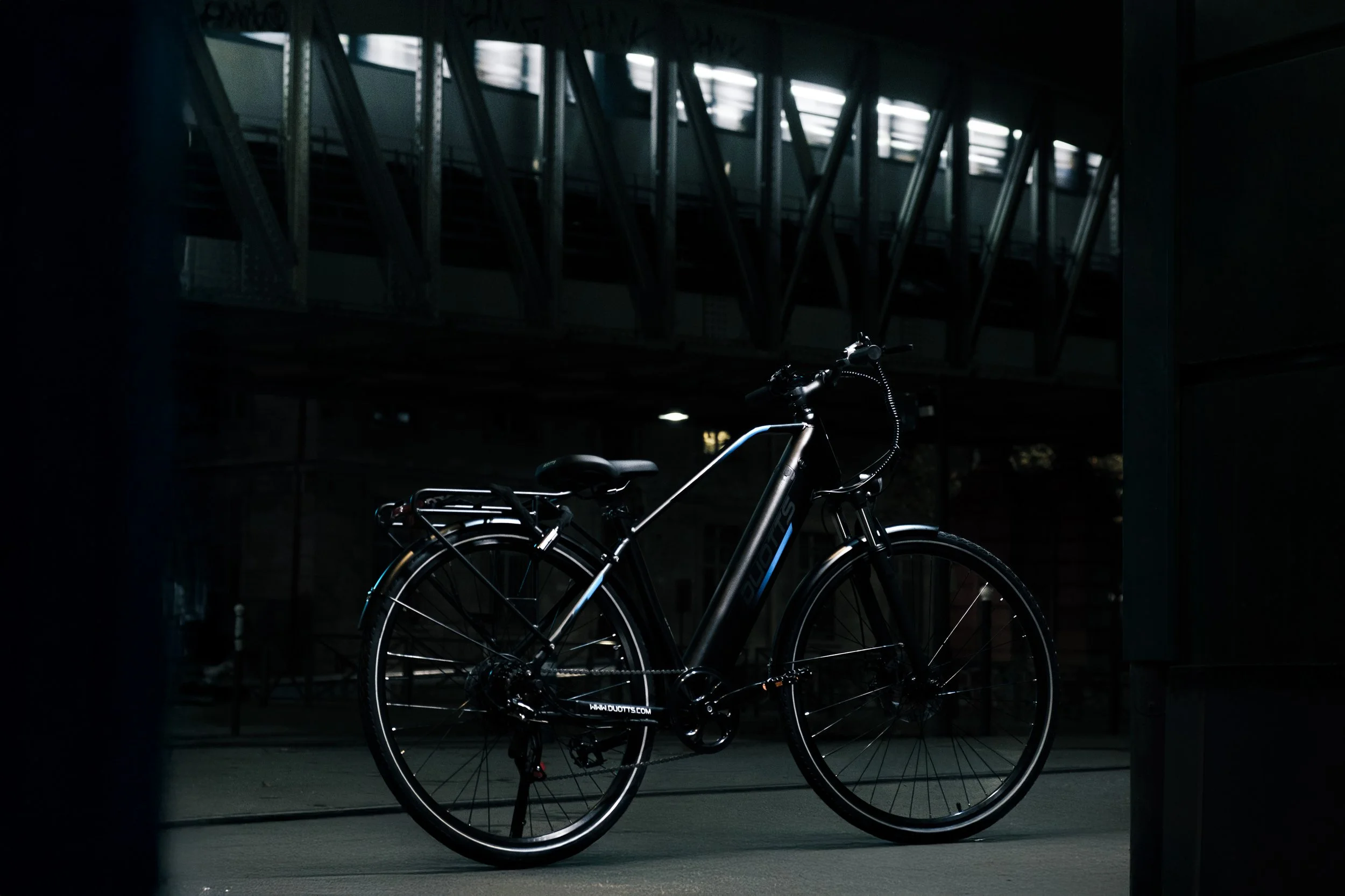 A black pedal bicycle with a rear rack is parked under a bridge at night, with the bridge's metal structure and windows in the background.