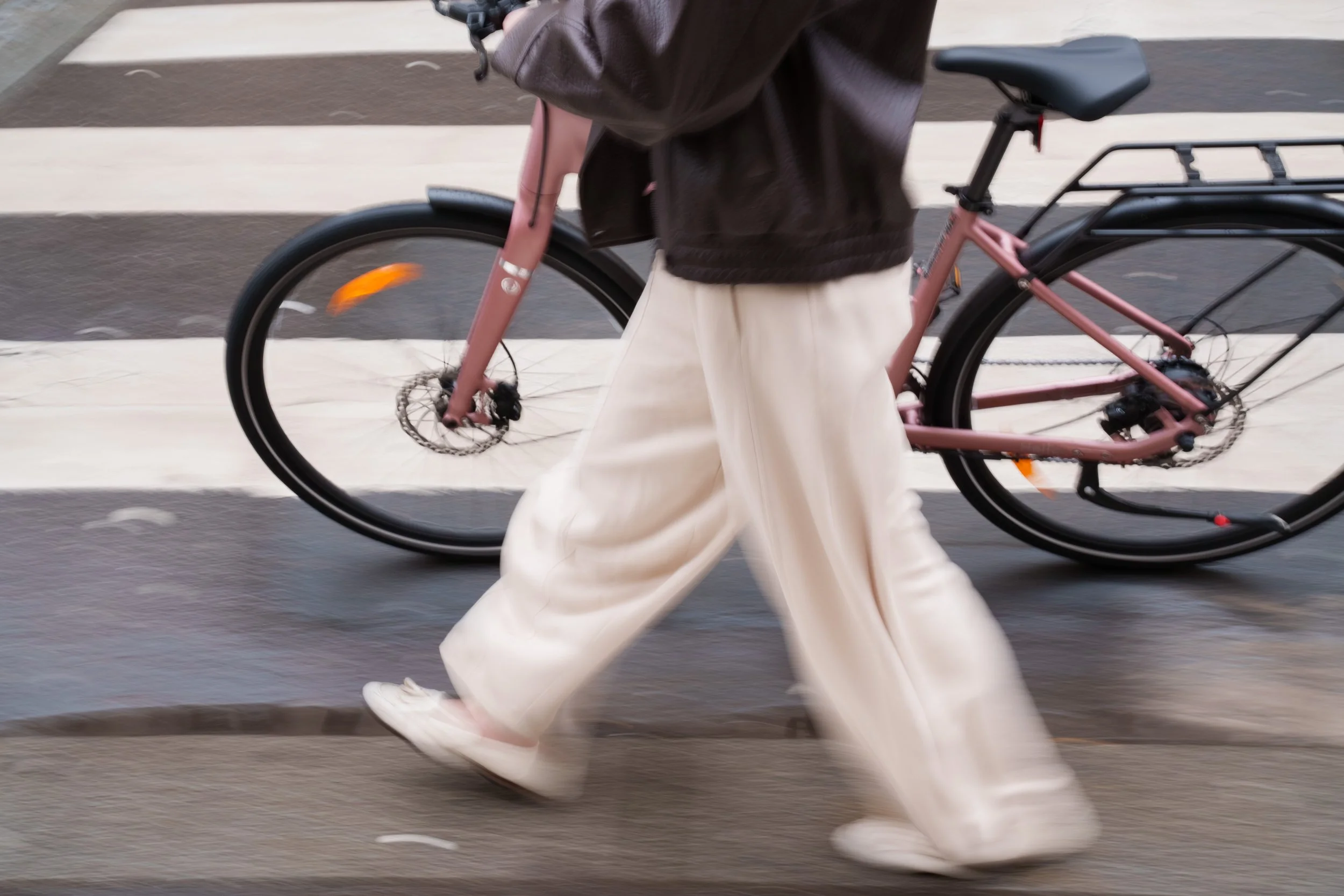 Person walking on a city sidewalk next to a pink bicycle with a rear rack, wearing white pants, a black jacket, and white shoes.