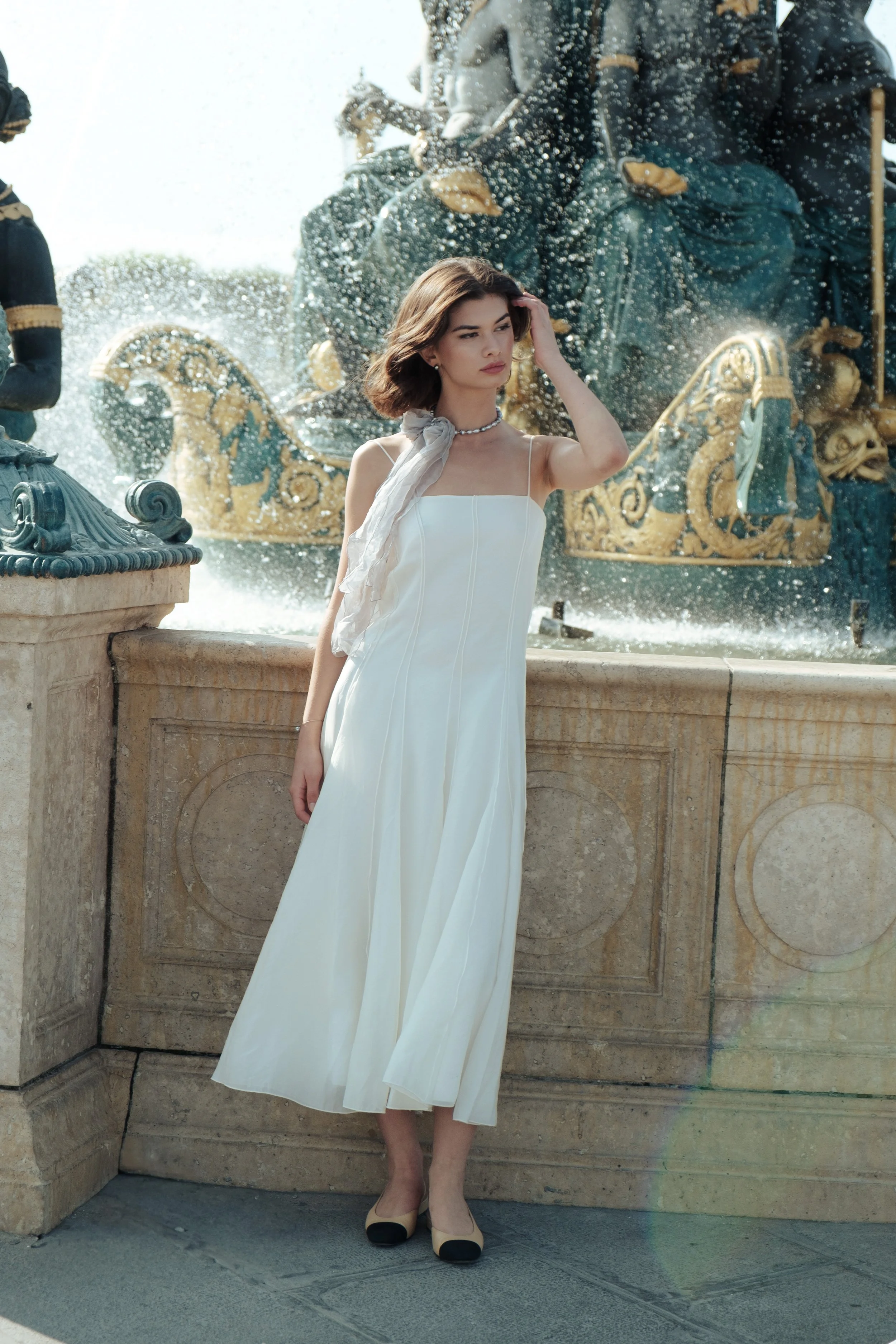 A woman in a white dress and black-and-cream heels standing near a fountain with a large ornate statue of a chariot and horses in the background.