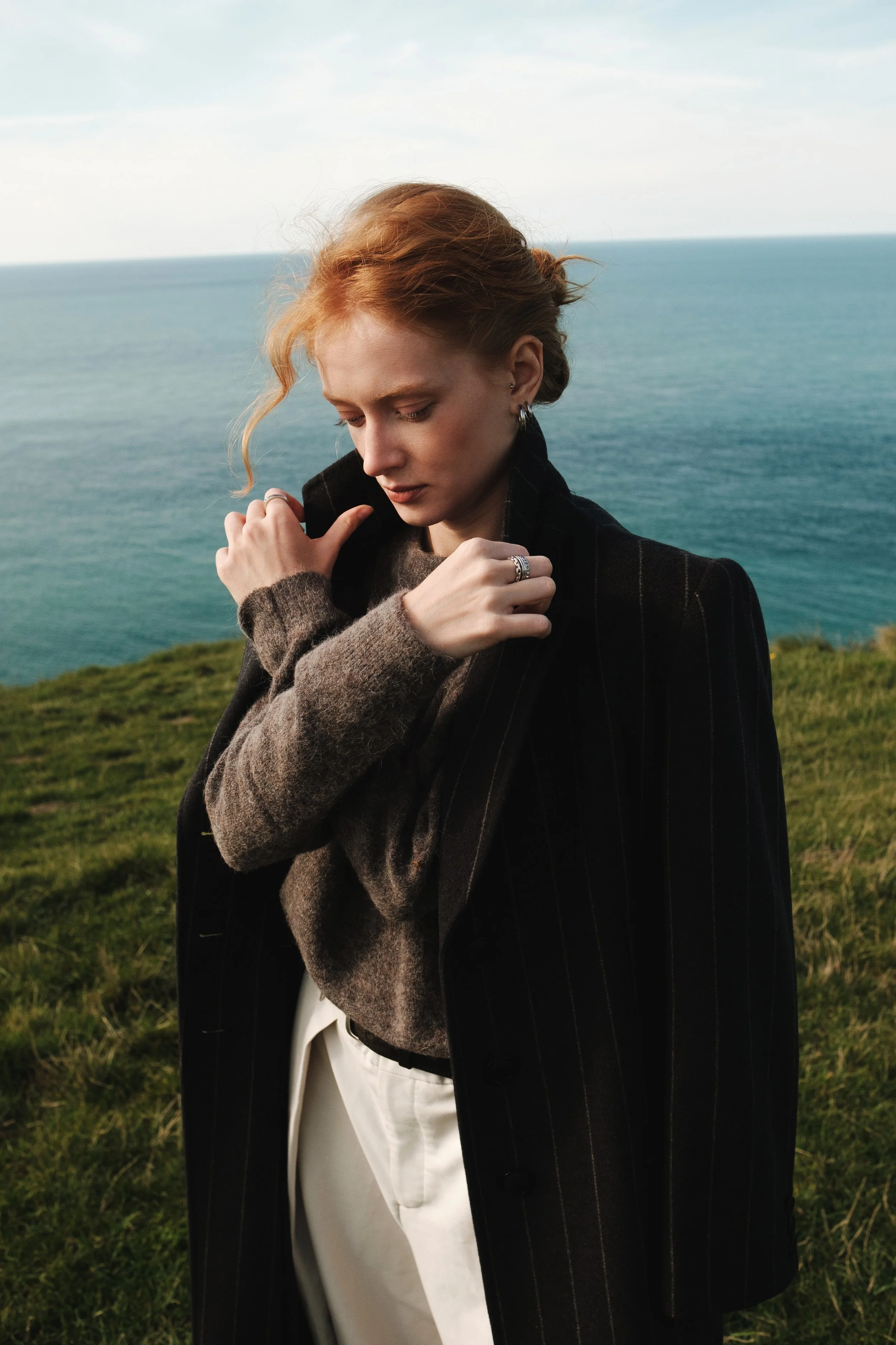 A woman with red hair standing on a grassy hill overlooking the ocean, wearing a dark coat and light-colored pants.