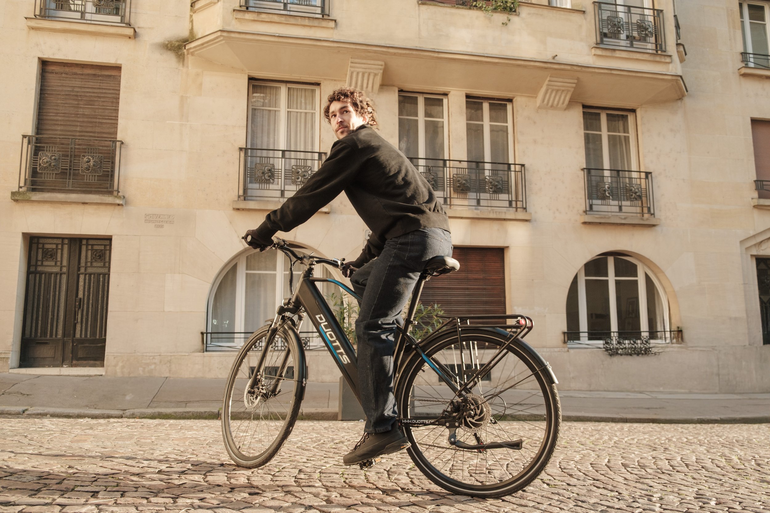 A man riding a bicycle on a cobblestone street in front of a beige multi-story building with balconies.