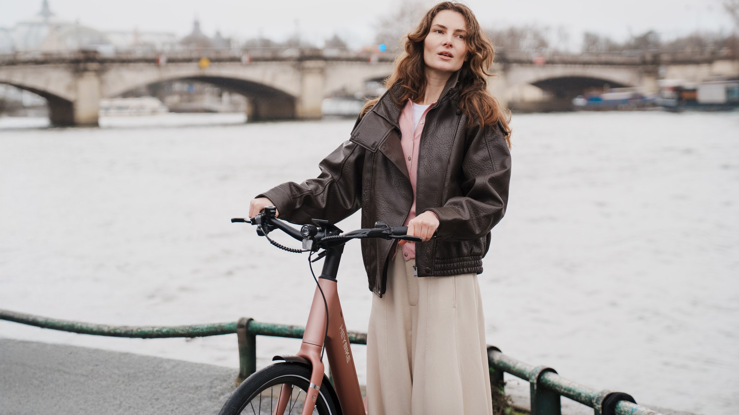 A woman with wavy brown hair standing next to a pink electric bicycle by a river, with a bridge and boats in the background.