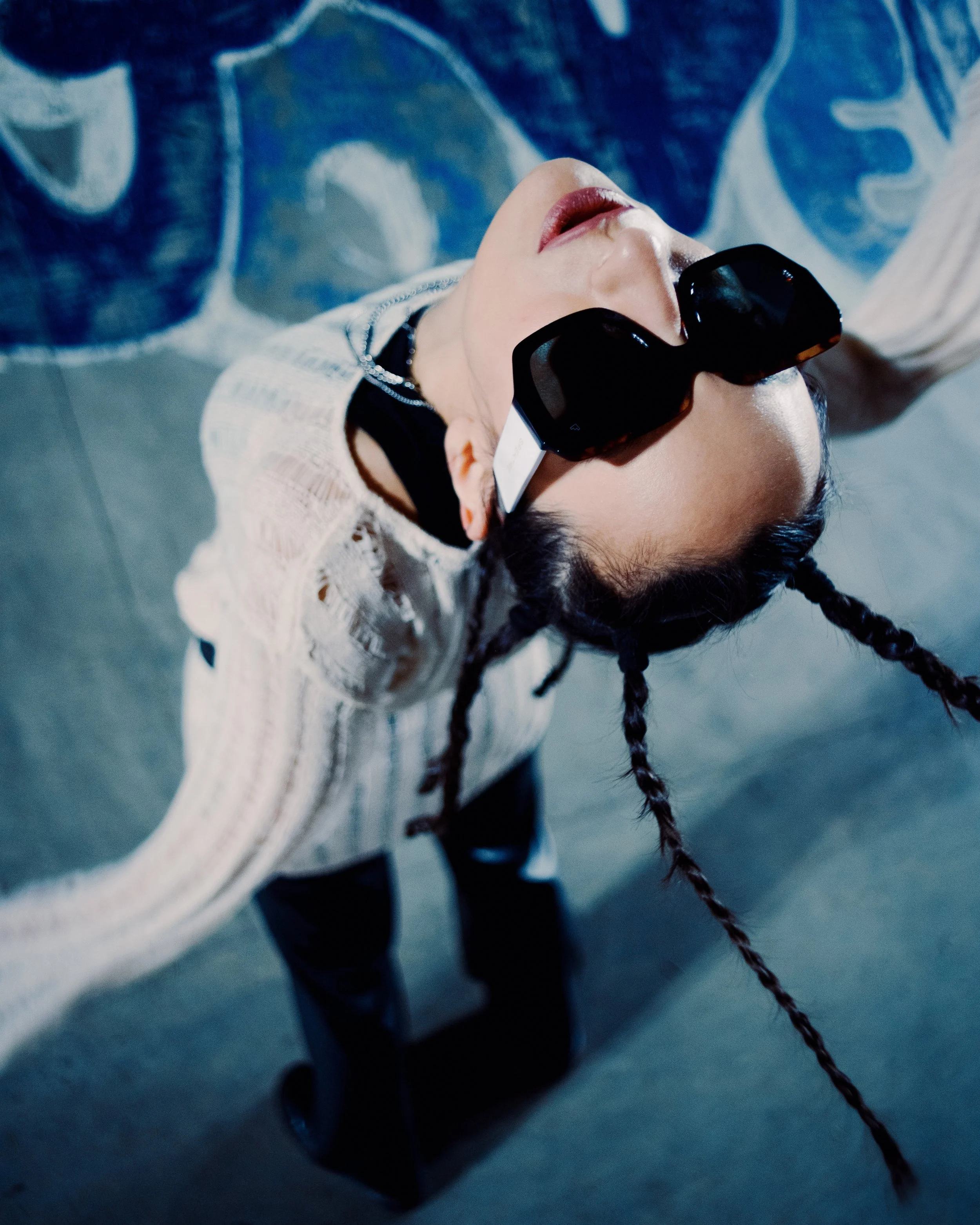 A woman with braided hair wearing large black sunglasses, a white textured top, and black pants, taking a selfie from above on a gray surface.