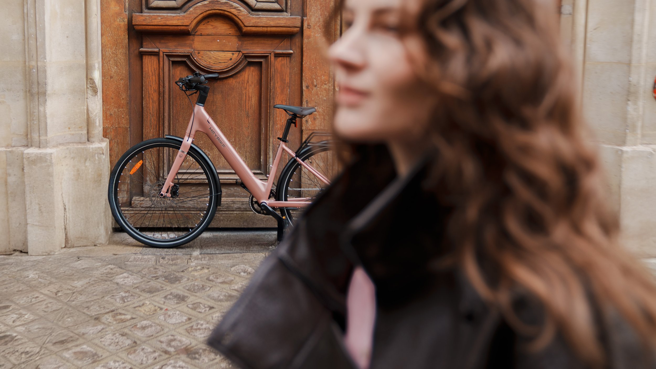 A woman with curly hair, wearing a dark jacket, standing outdoors near a stone building with a wooden door, with a pink bicycle parked against the door in the background.