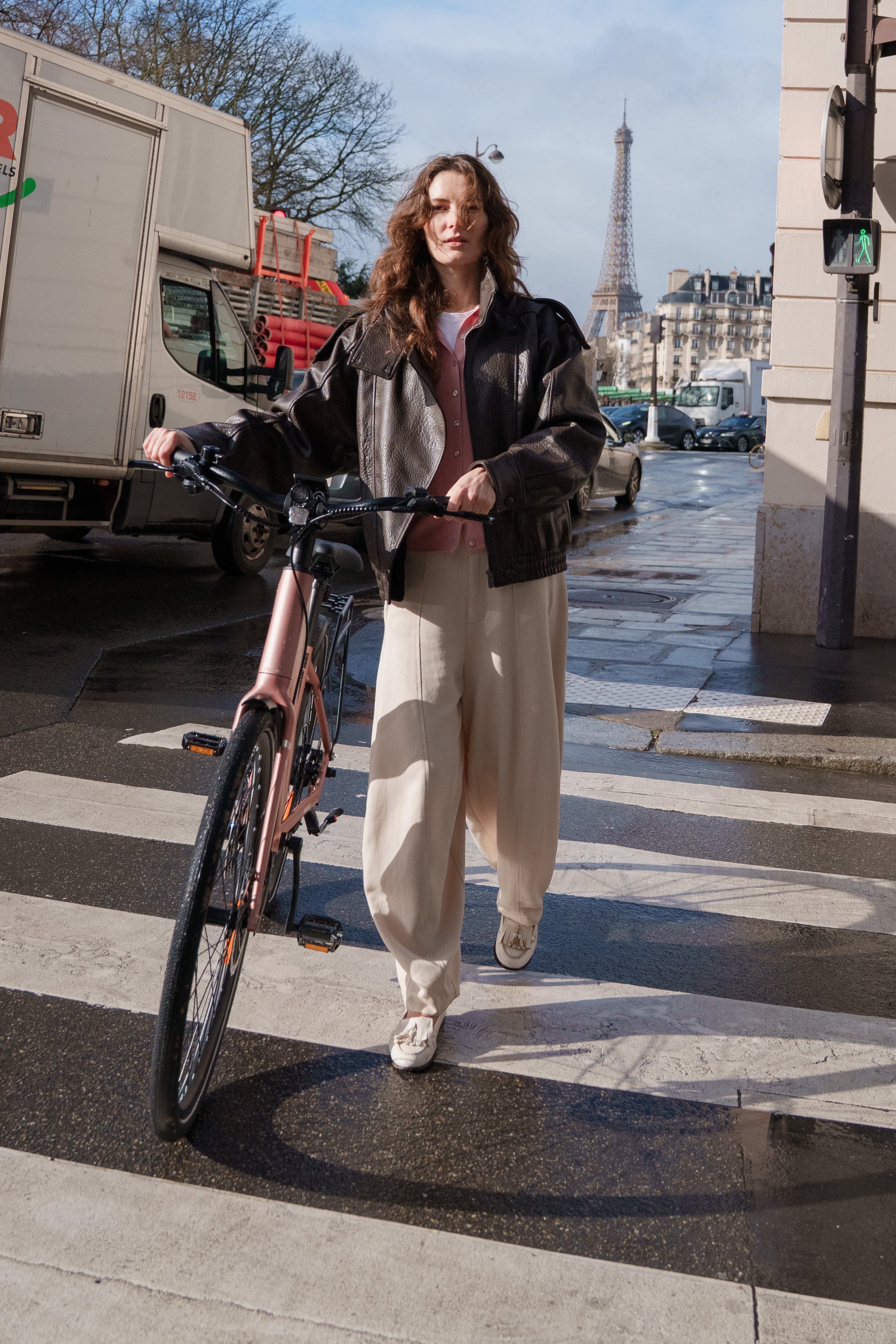 A woman with wavy brown hair crossing a street in Paris, holding a pink bicycle, with the Eiffel Tower visible in the background. She is wearing a brown leather jacket, pink shirt, white pants, and white shoes.