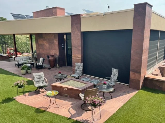 Outdoor patio with four chairs, two small side tables, a custom stone outdoor fire pit, and potted plants, adjacent to a modern house with large glass windows, a closed black garage door, and a custom stone exterior by Boulder StoneScapes.
