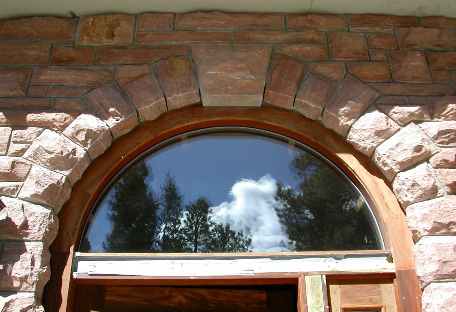 Close-up of an arched window with a wooden frame set in a custom stone wall built by Boulder StoneScapes out of Boulder, CO reflecting a sky with clouds and trees.