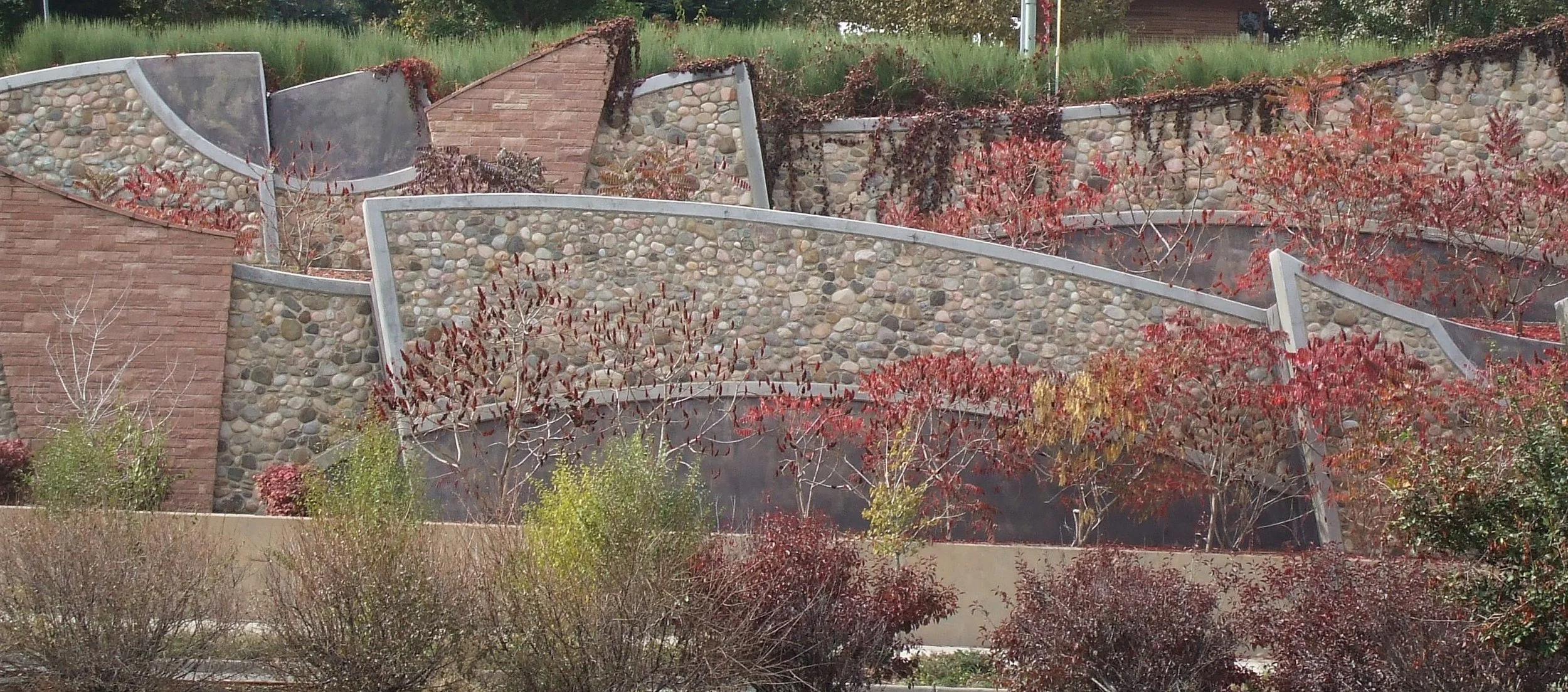A custom stone cityscape outdoor area with a curved stone wall and metal accents, accented by various plants and red foliage created and built by Boulder StoneScapes.