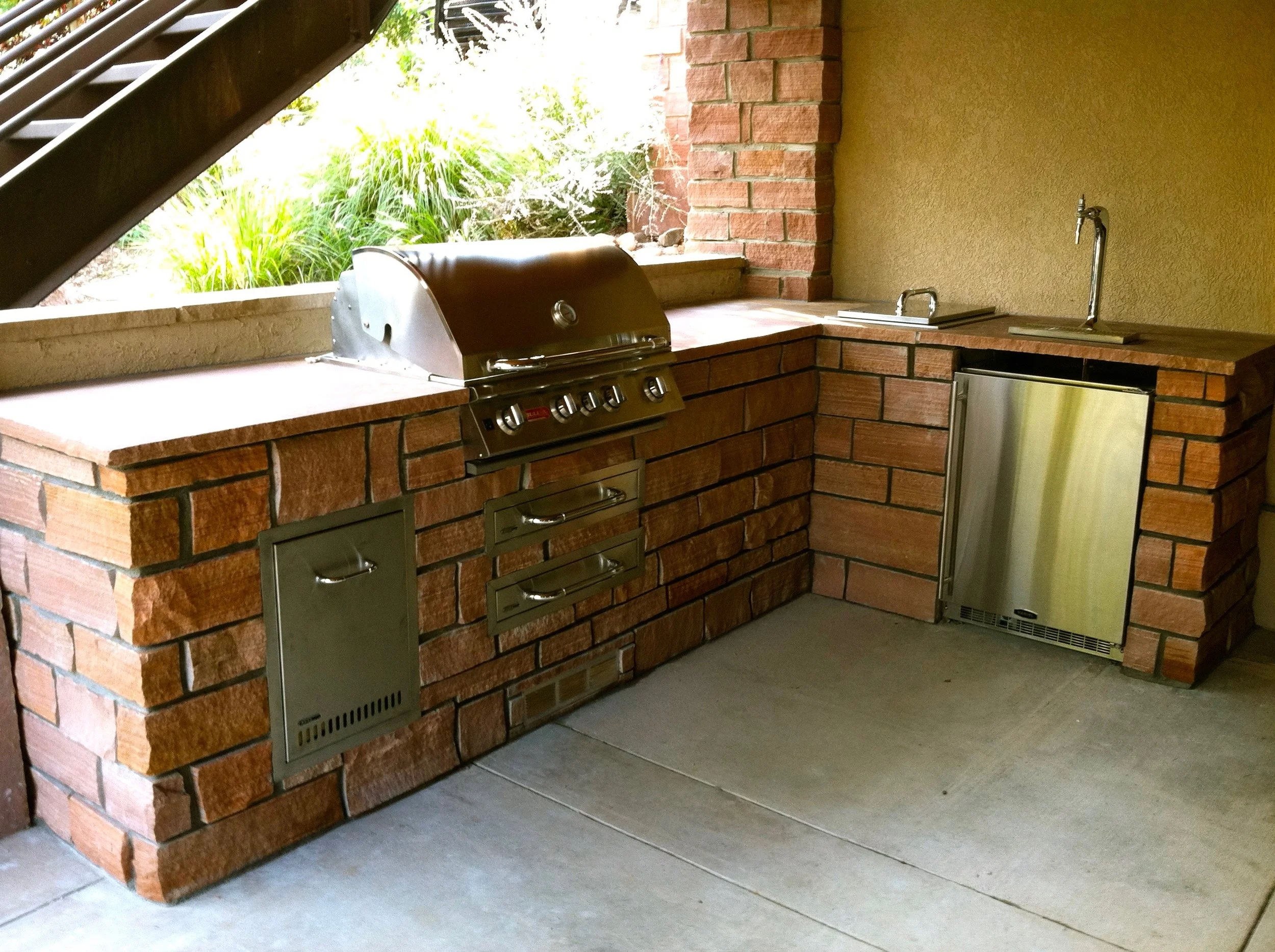 Outdoor kitchen with brick counter, custom stone work, built-in grill, small stainless steel refrigerator, and sink, under a covered area near stairs and greenery