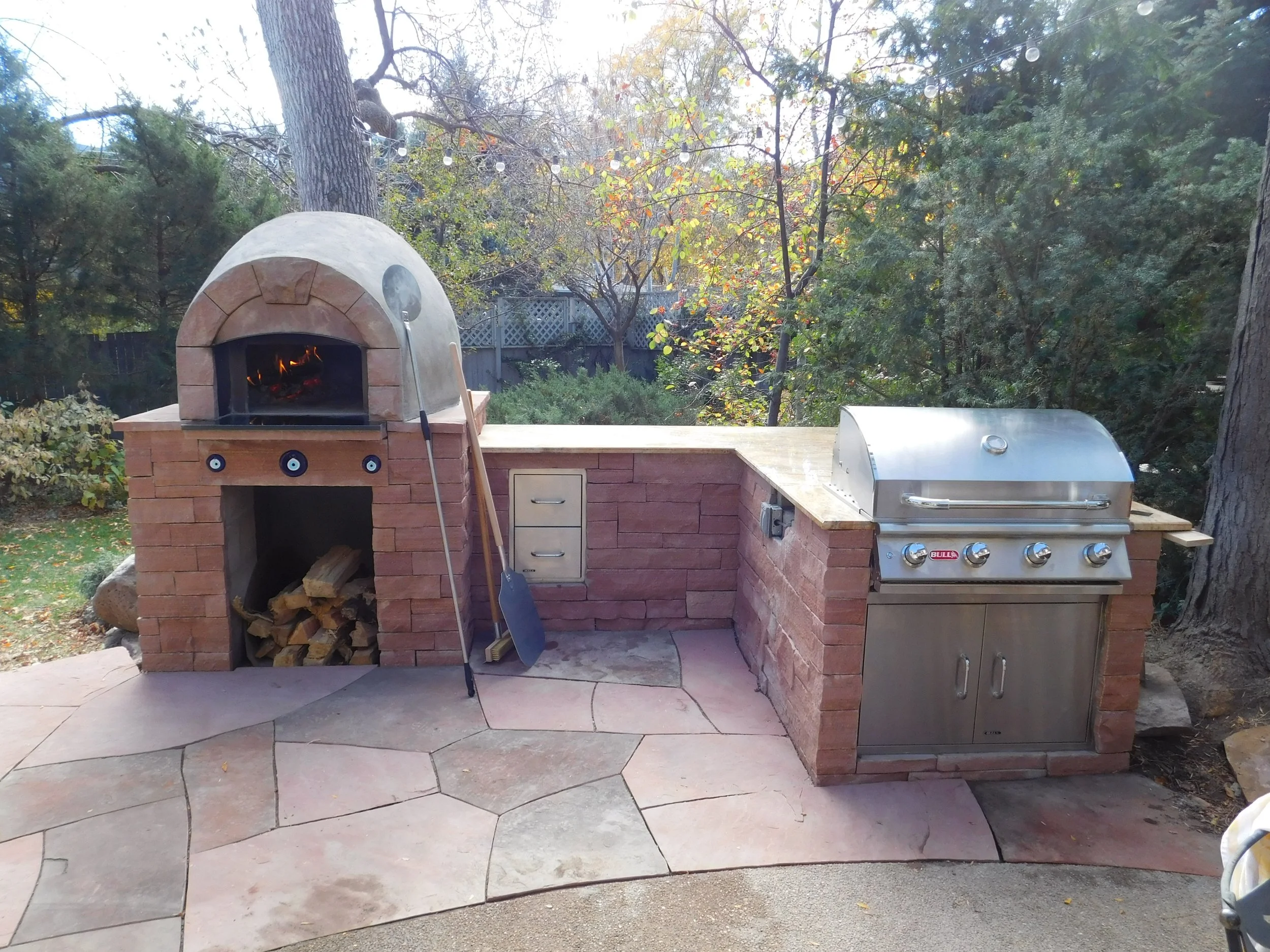 An outdoor kitchen setup with a brick pizza oven with a chimney, stacked firewood inside, and a metal gas grill with four control knobs and a closed lid, all set on a stone patio with trees and foliage in the background.