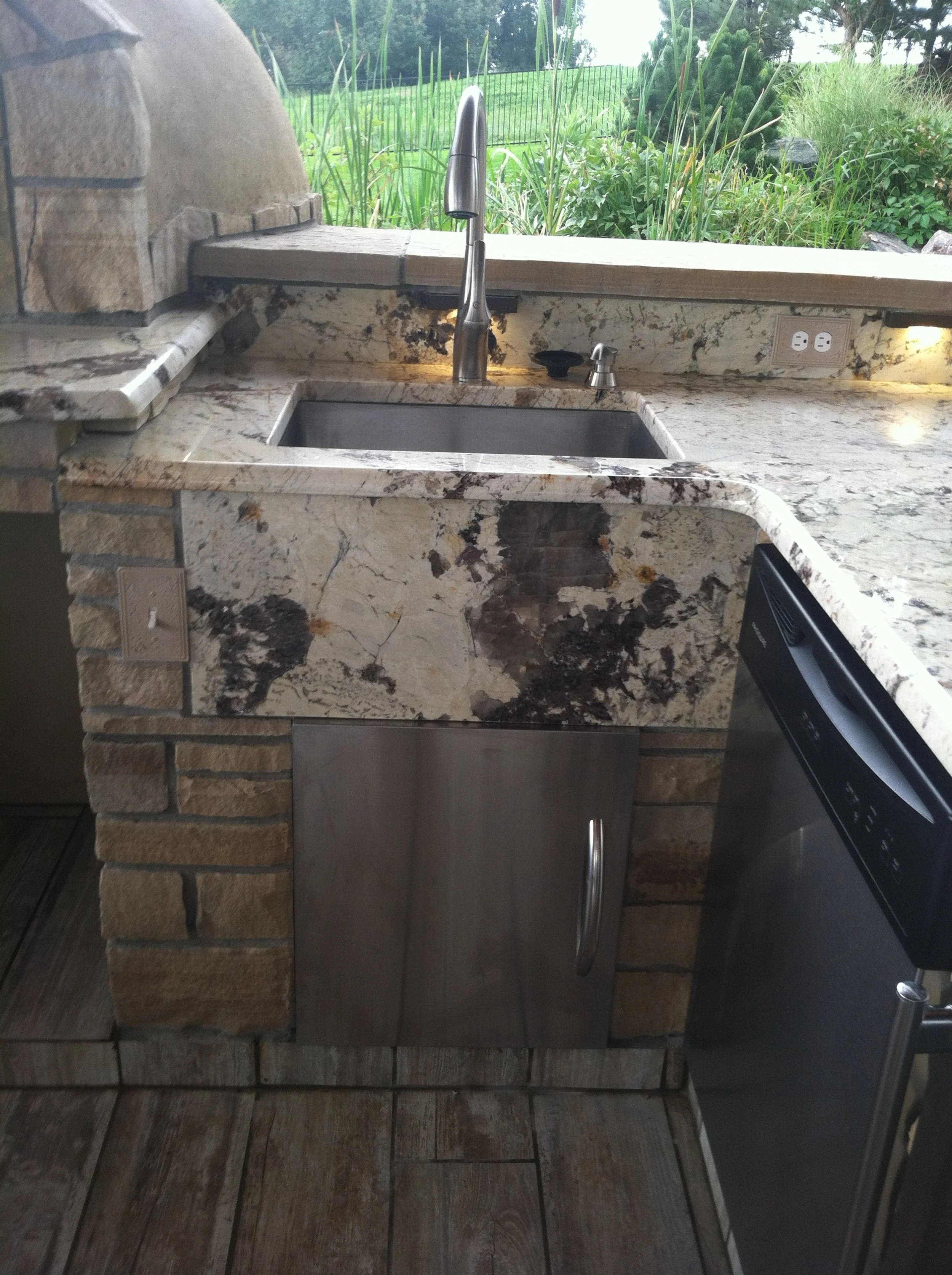 Outdoor kitchen with a marble counter, stainless steel sink, and faucet. There is a patch of greenery outside, electrical outlets, and a dishwasher below the counter.