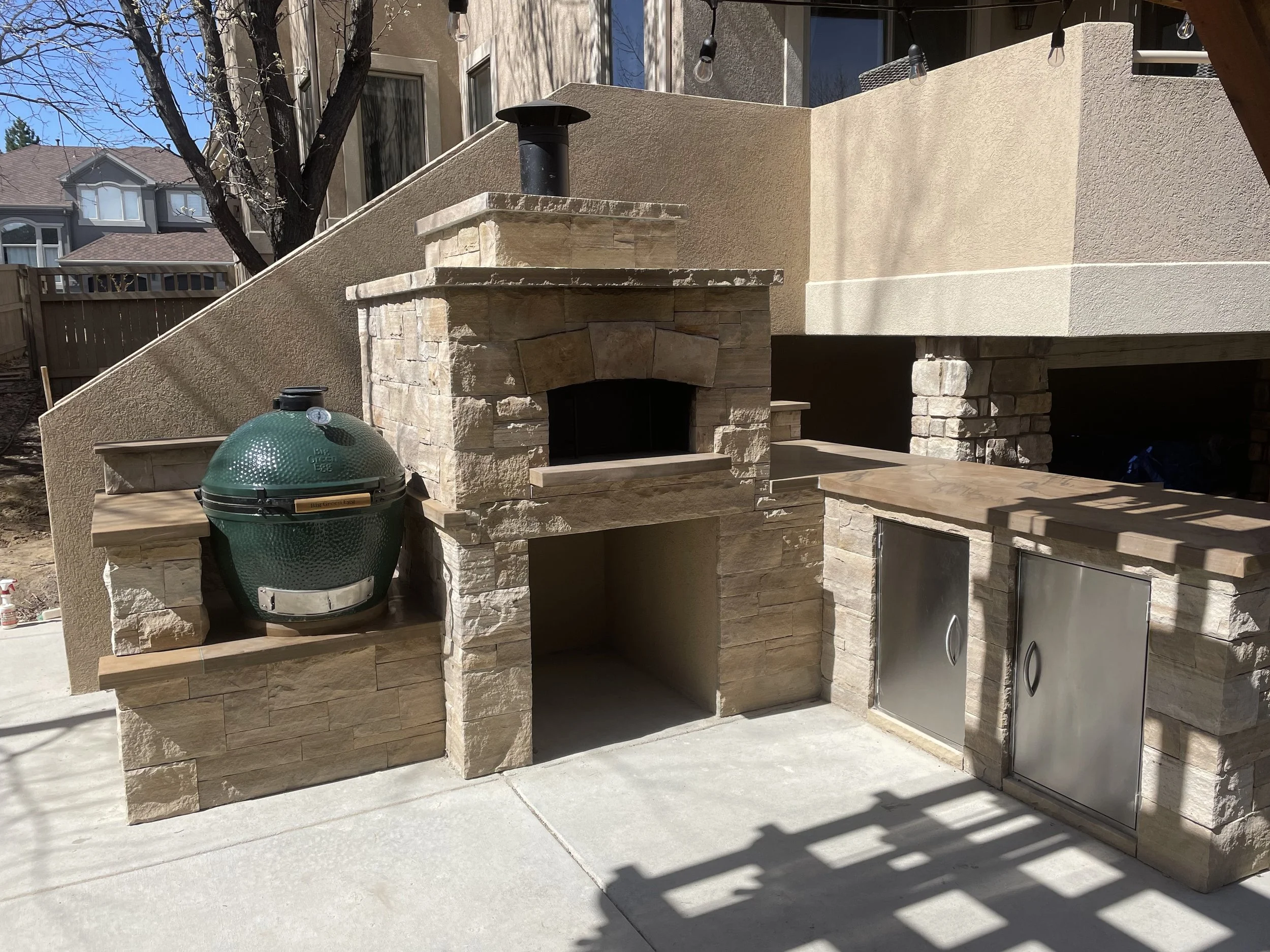 Outdoor kitchen area with a built-in stone pizza oven, a green ceramic barbecue grill, and stainless steel storage cabinets, with beige and stone finishes, under a raised patio with string lights.