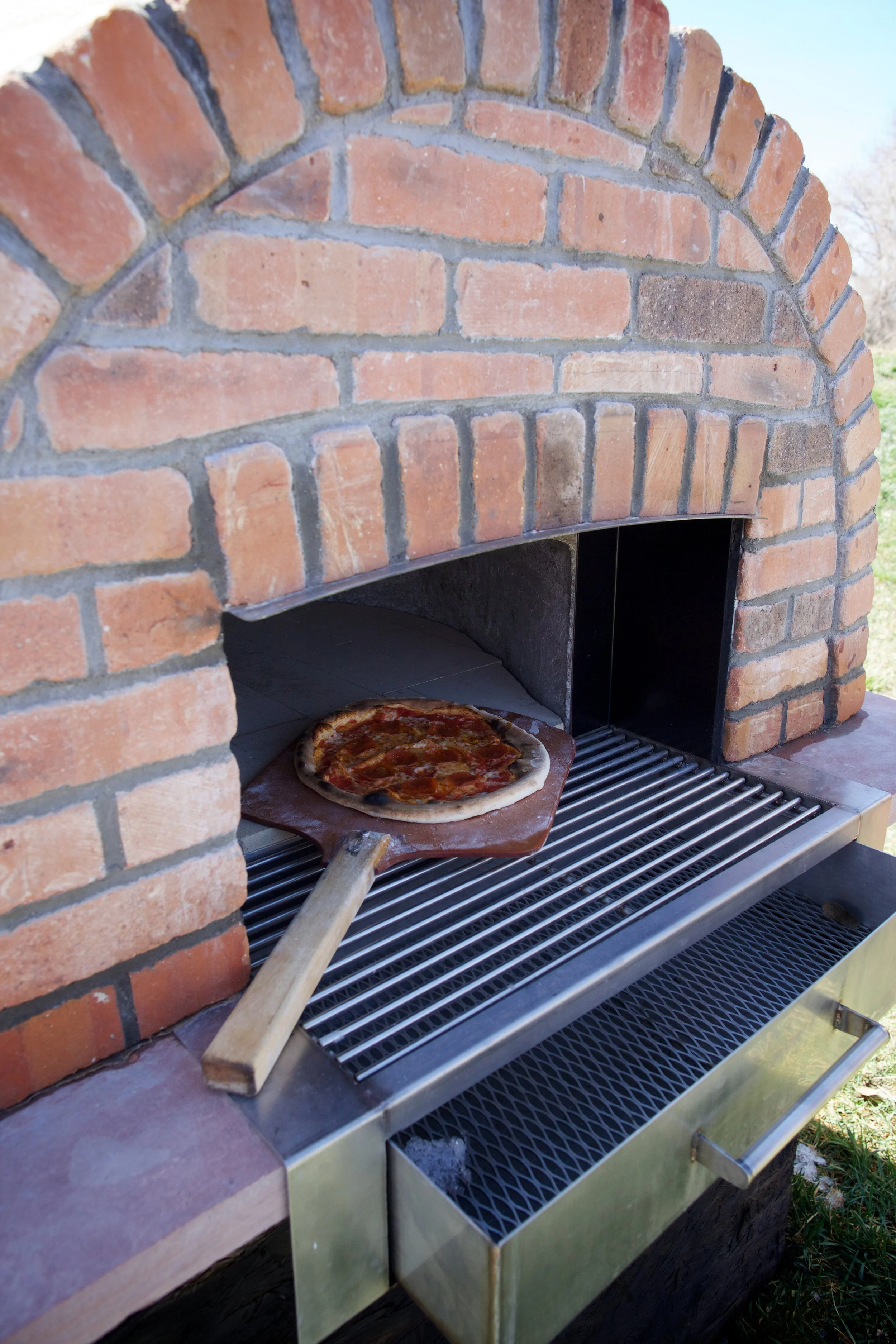 A custom stone outdoor pizza oven with a pizza peel holding a pizza inside built by expert stone masonry Boulder StoneScapes.