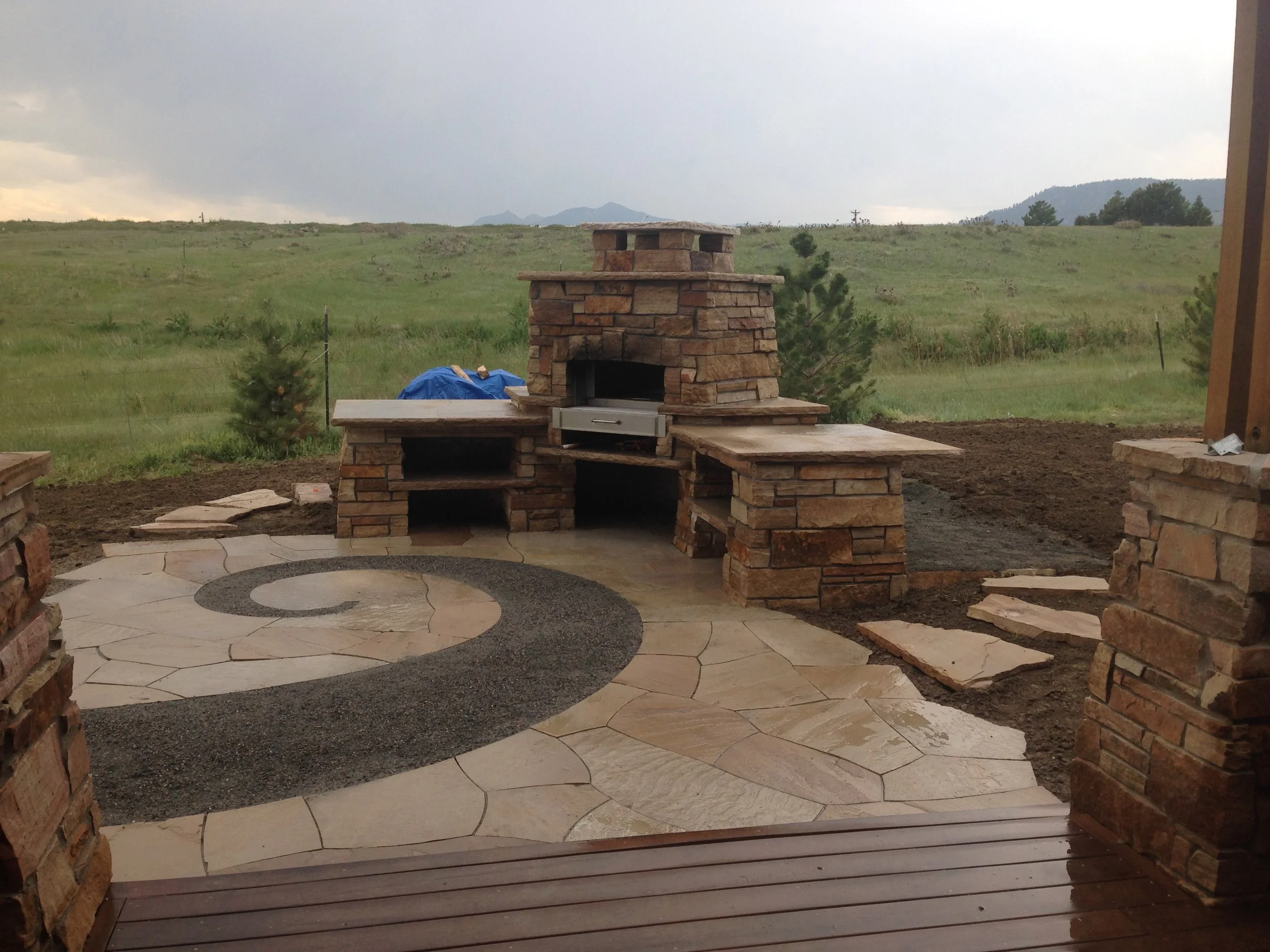 Outdoor stone fireplace and kitchen with stone countertops, surrounded by a paved patio with a spiral design, overlooking a grassy field and mountains in the distance under a cloudy sky.