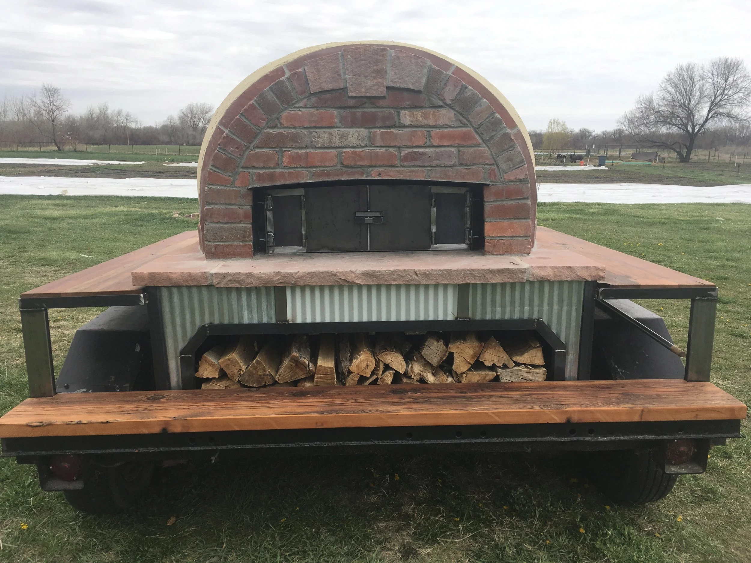Wood-fired outdoor oven with a stone masonry dome, a wood storage compartment below, and a wooden work surface on each side, situated on a trailer on a grassy field.