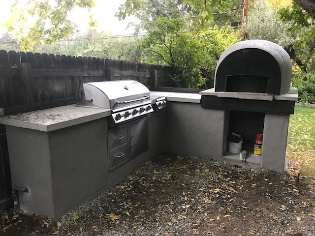 Outdoor stone kitchen with a modern grill and a built-in pizza oven under a tree in a backyard.