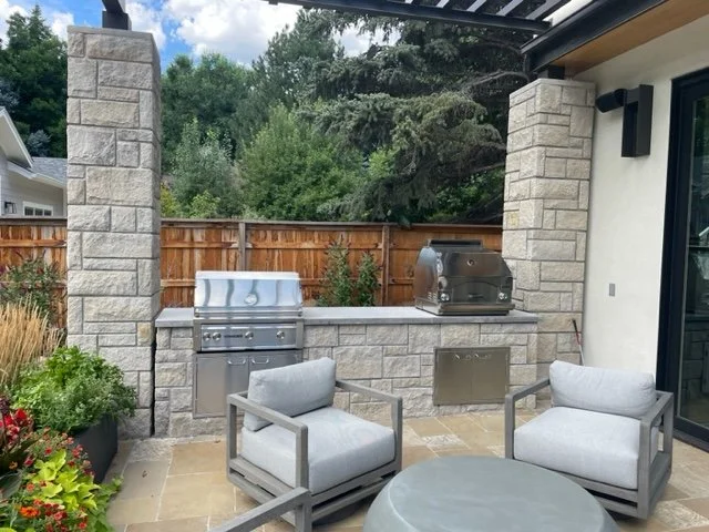 Outdoor patio with two gray cushioned chairs, a round gray table, a stainless steel grill, and a wooden fence in the background with custom stone work by Boulder StoneScapes.