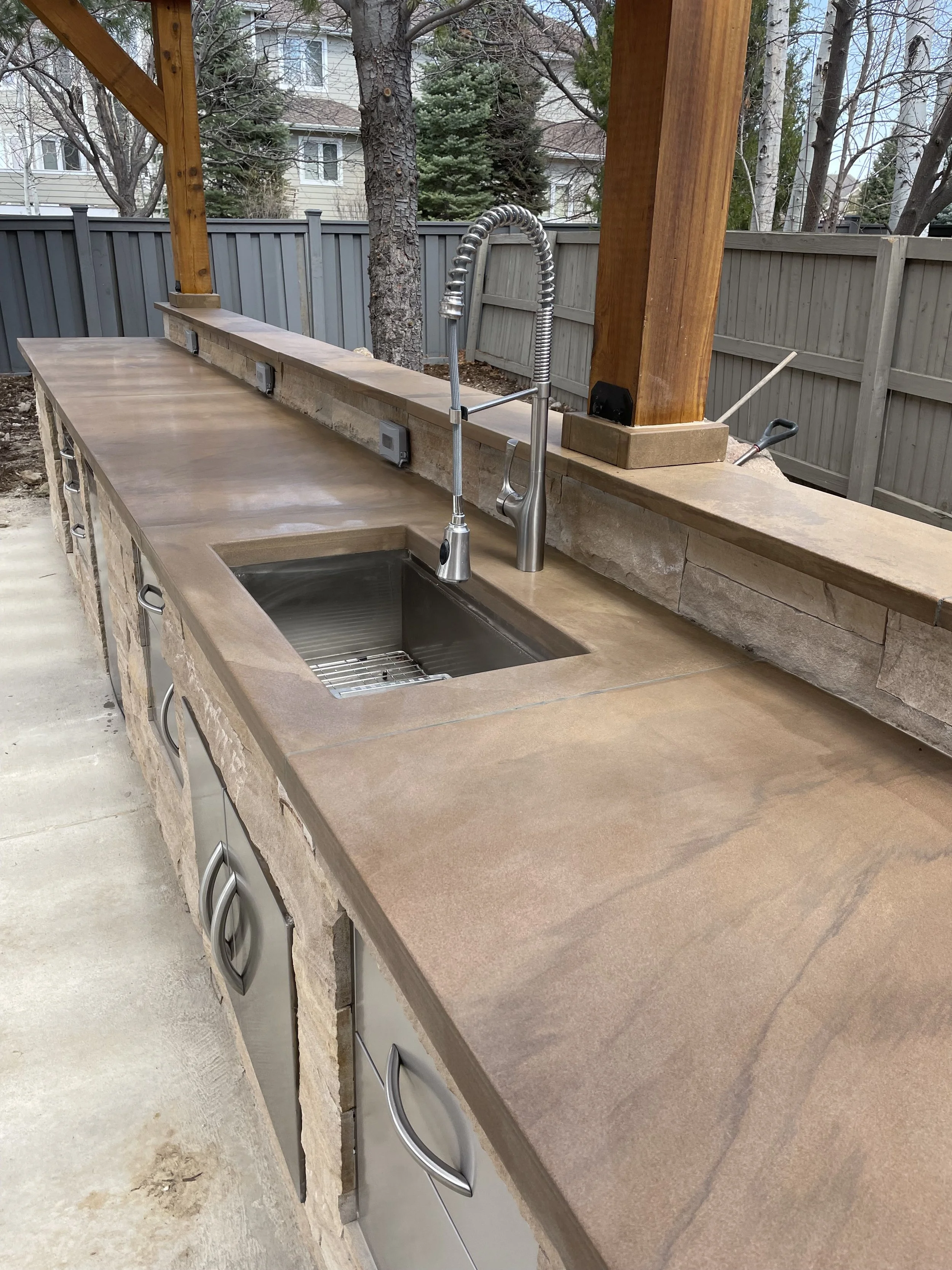 Custom outdoor kitchen with a brown stone countertop, stainless steel sink, and a high-arc faucet. The kitchen is built with stone and wood materials, with a wooden post and a raised stone bar in the background, and a gray fence.