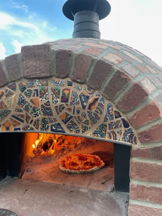 A custom stone masonry brick pizza oven with colorful mosaic tiles around its arch, burning fire inside, with a pizza on the oven floor crafted by Boulder StoneScapes.