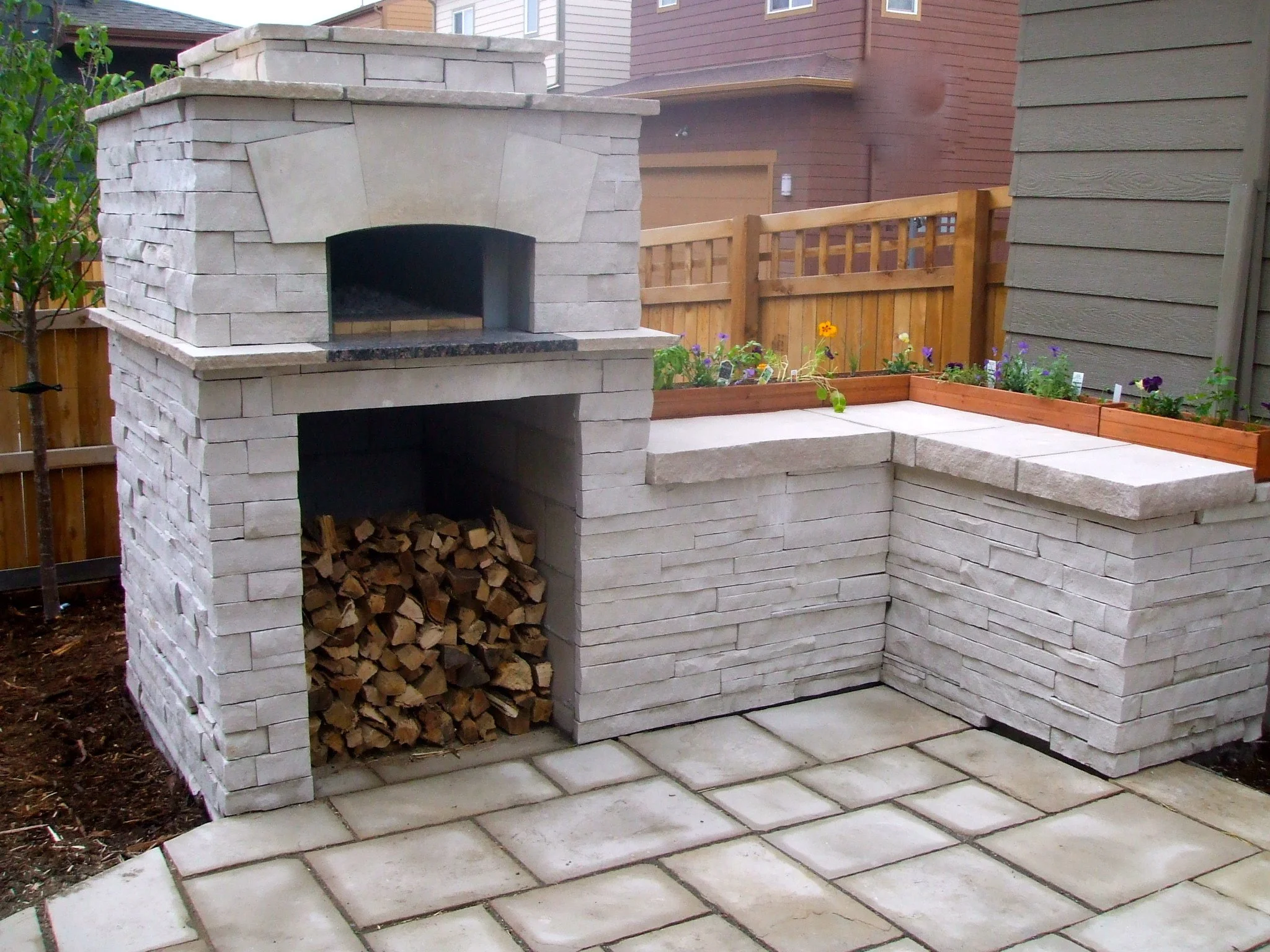 An outdoor custom stone fireplace with stacked firewood underneath, next to a garden bed with colorful flowers and a wooden fence in the background.